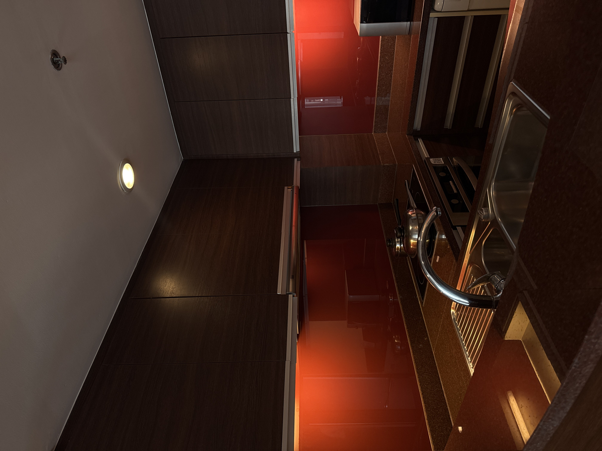 The image shows a modern kitchen with dark wood cabinets and a vibrant red backsplash. The countertops appear to be a speckled dark granite. In the foreground, a stainless steel sink with a gooseneck faucet is visible, along with a dish rack. A stovetop with pots and pans is situated behind the sink. On the right side of the frame, a silver microwave is partially visible. The overall lighting is dim, with an overhead light illuminating the ceiling and a warm glow emanating from under the cabinets. The scene suggests an interior residential setting. The location is identified as Bangkok, Thailand. There are no people or animals present in the image.
