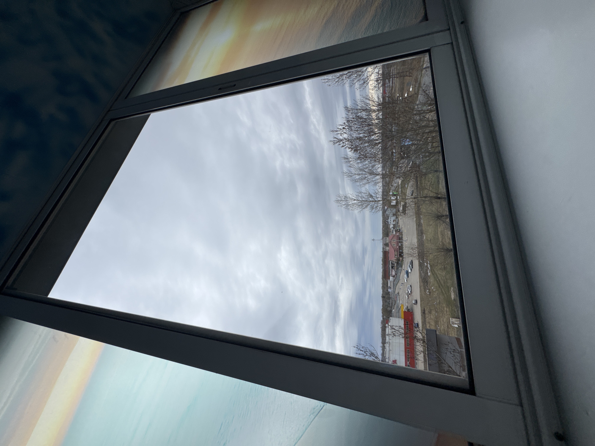 A view through a window. The window frame is grey and the glass pane shows a cloudy sky above a commercial area in Kladovitsy, Russia. A parking lot contains many cars and there is a large building with a red roof and a yellow accent. Bare trees are visible in the distance, along with other buildings and vegetation. The interior wall adjacent to the window is white. Wallpaper with a sky or water theme is visible on the other adjacent walls.