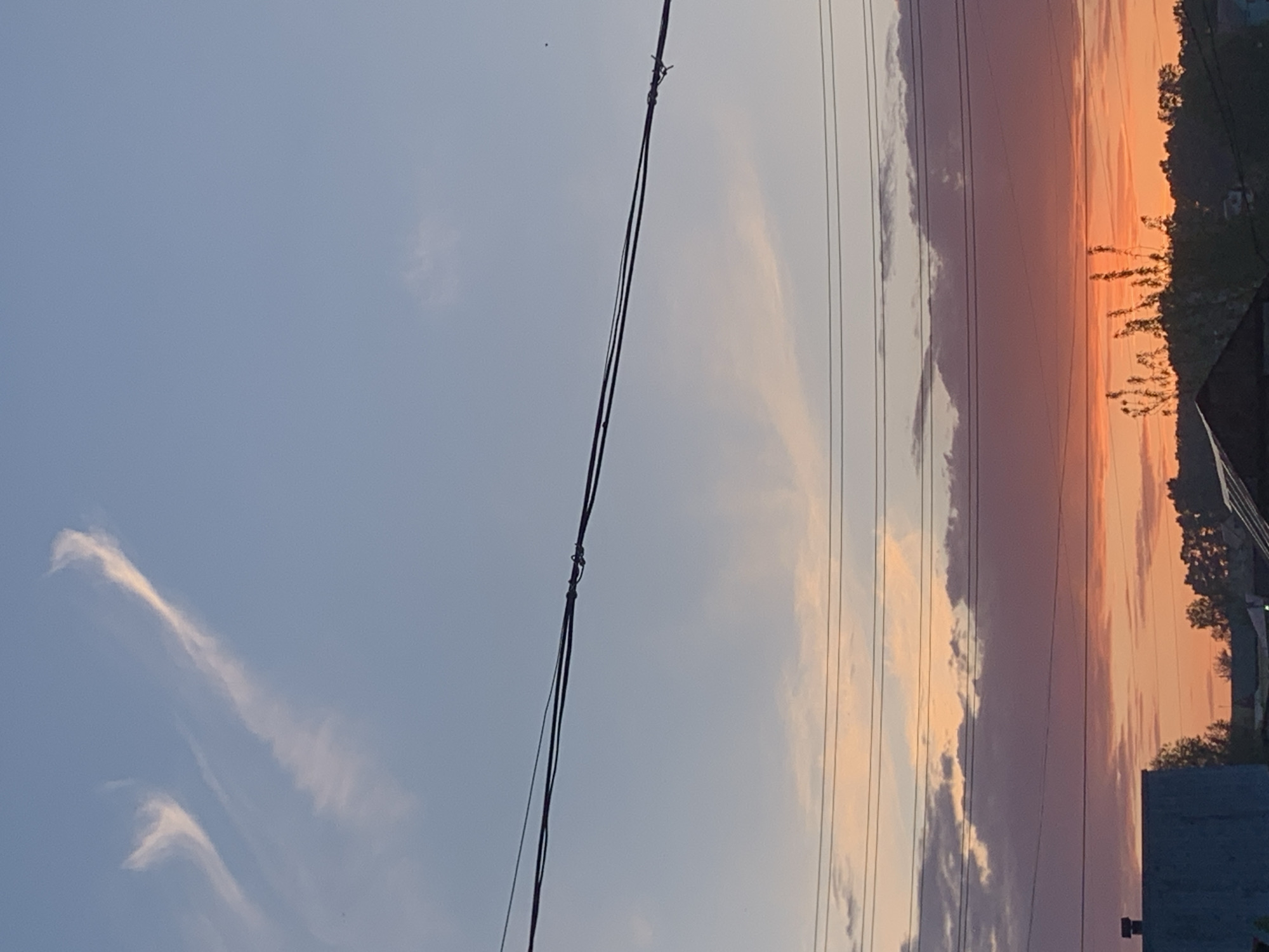 The image depicts a view in Фоки, Russia, likely at sunset or sunrise. The sky is transitioning from a light blue to hues of orange and pink, with clouds streaked horizontally across the view. Power lines are prominently visible in the foreground, intersecting the sky. Trees and rooftops of buildings are visible along the horizon.