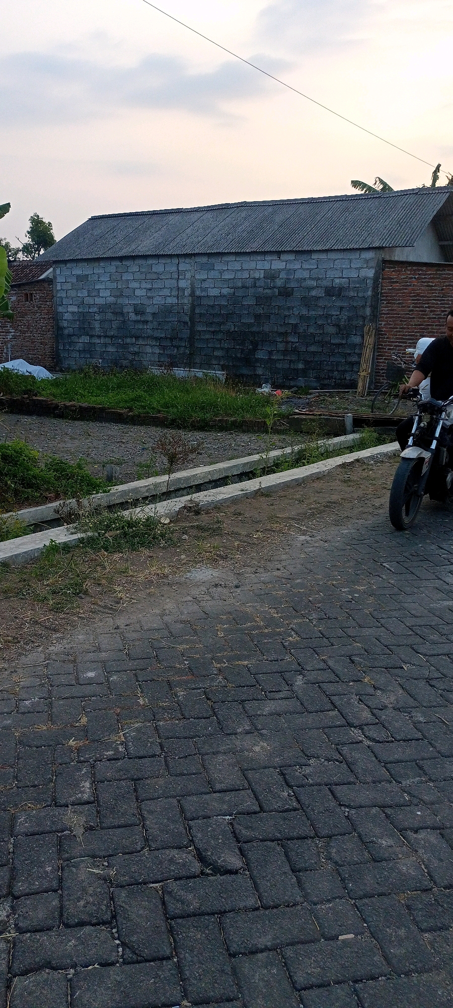 In Gempolklutuk, Indonesia, a man is riding a motorcycle on a paved brick road. The motorcycle is white and black, and the man is wearing a black shirt. In the background, there is a long building with a gray corrugated metal roof and cinder block walls. The building appears to be under construction or renovation. There is also some vegetation and a drainage ditch near the building. The sky is cloudy and overcast.