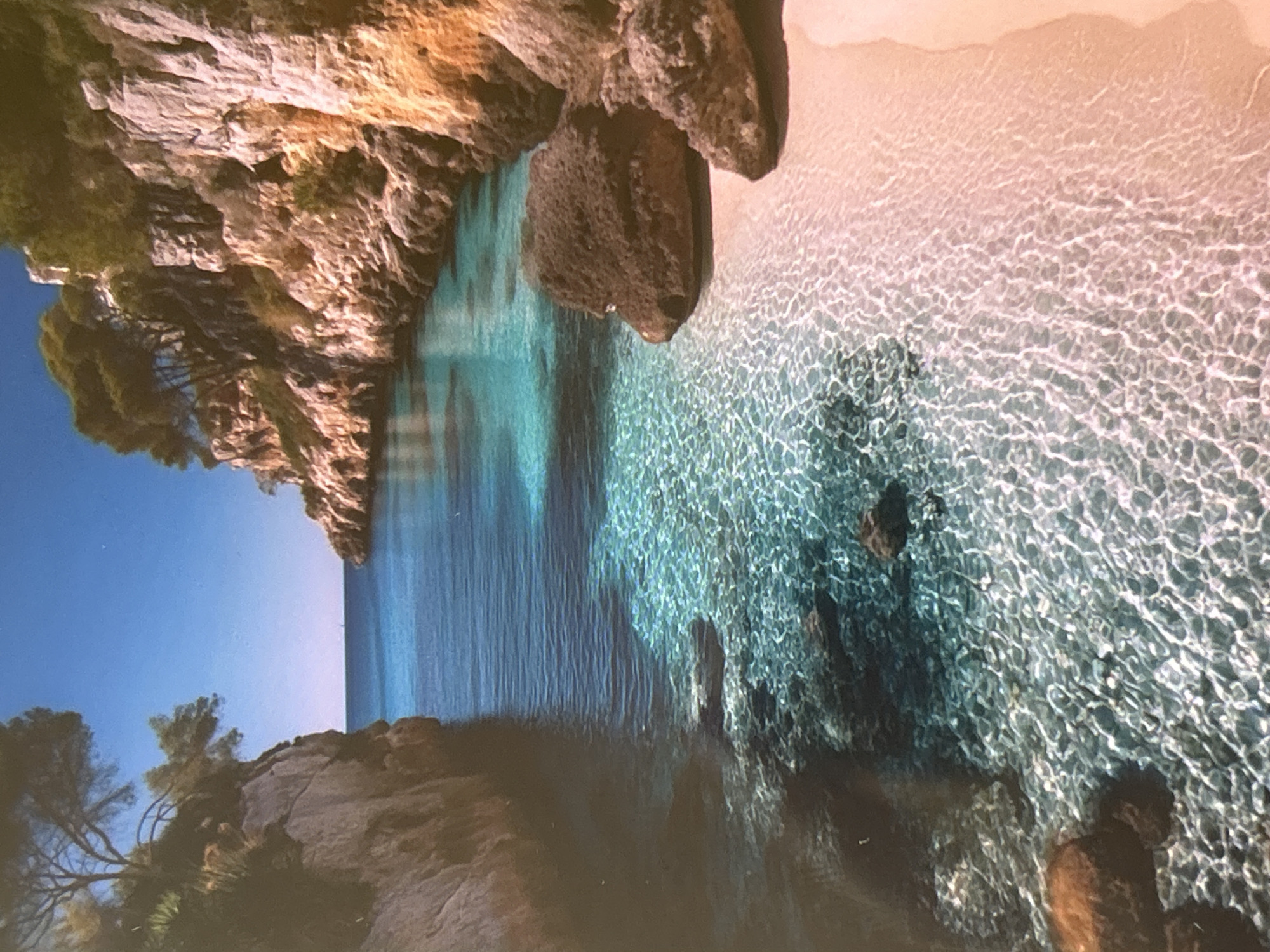 This image captures a breathtaking, elevated view of a secluded coastal cove under clear, sunny skies. The dominant features are the exceptionally clear, vibrant turquoise waters and the rugged, rocky cliffs that frame the inlet.
On the left side, a tall, textured cliff face, displaying hues of brown, orange, and gray, rises steeply from the water. It is sparsely covered with green trees and vegetation, clinging to its ledges and crevices. Another smaller, darker rocky formation extends into the water from the upper right, almost meeting the main cliff face, creating a narrow entrance to the cove.
The water exhibits a stunning clarity, especially in the shallower areas closer to the shore on the right. Sunlight penetrates the surface, creating intricate, shimmering patterns of light and shadow on the sandy bottom, which appears a light reddish-beige. As the water deepens towards the center and left, it transitions to a profound, dark blue, suggesting significant depth. Several dark, submerged rocks are visible beneath the pristine surface.
A segment of a sandy beach, matching the light reddish-beige color seen through the water, is visible along the far right side of the image, curving gently away from the clear waters. The sky, visible in the upper left quadrant, is a serene, unbroken blue, completing the idyllic natural landscape. No people, man-made objects, or visible text are present, emphasizing the untouched beauty and tranquility of the scene.