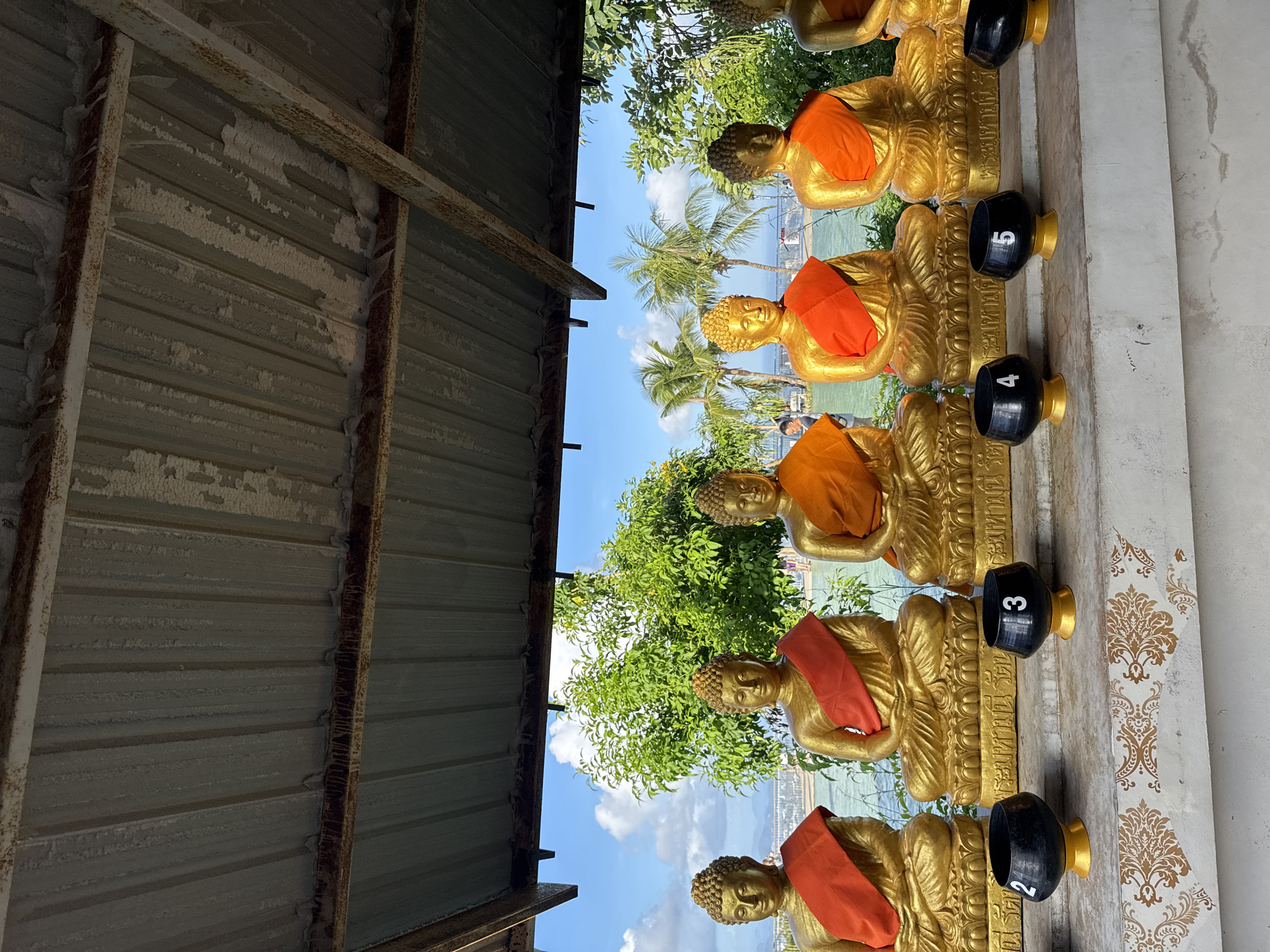 The image captures a row of seven golden Buddha statues adorned with orange sashes, arranged under a corrugated metal roof. Each statue sits on a raised platform inscribed with Thai script. In front of the statues, a line of black alms bowls, some marked with numbers "2", "3", "4", and "5", are placed on a decorative ledge. The background reveals a picturesque scene of lush green trees, palm trees, and a body of water under a bright blue sky with scattered clouds, indicative of a tropical coastal environment, likely in Karon, Thailand. The overall ambiance suggests a peaceful and serene religious or cultural site.