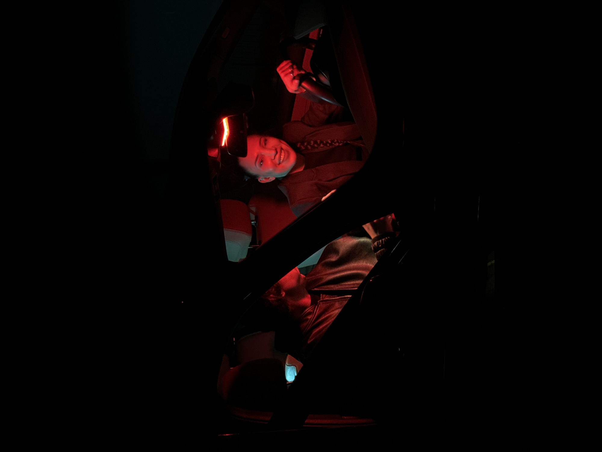 Two individuals are visible inside a vehicle, illuminated by red and blue lights. A woman, wearing a jacket or uniform and a braided hairstyle, is smiling and gripping the steering wheel. Below and partially obscured by the car's interior, a man wearing a leather jacket is visible, looking down at a lit screen of a mobile device. The scene is very dark, with the interior of the car and the subjects within it being the primary areas of illumination. The location is not identifiable as Moscow, Russia based on the visual data provided.