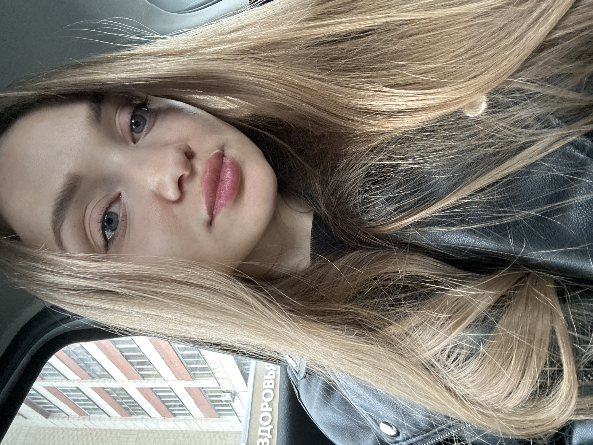 The image features a close-up selfie of a young woman with long, blonde hair. She has fair skin, blue eyes, and pink lips. She is wearing a black leather jacket. The background shows a building with a red and white striped facade. The Russian word "ЗДОРОВЬЯ" (meaning "health") is partially visible, suggesting the presence of a health-related facility. Based on the architectural style and the presence of the Russian word "ЗДОРОВЬЯ", the location is likely Kazan, Russia. The woman is possibly sitting inside a car, based on the angle and framing of the shot.