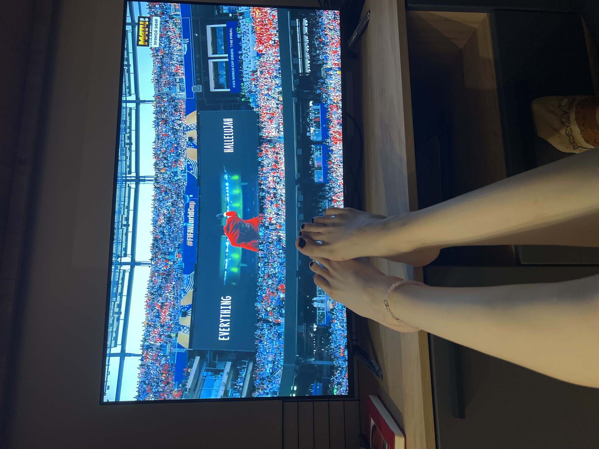 A television screen displays a scene from the FIFA World Cup 2026 final. The screen shows a stadium filled with spectators, a performer in a red outfit on stage, and text that reads "EVERYTHING," "#FIFACup," and "HALLELUJAH." A broadcast logo for "MATCH!" is visible in the top right corner of the screen. In the foreground, a person's legs and feet are visible, with one foot adorned with black nail polish and a delicate ankle bracelet. The legs are resting on a wooden surface in front of the television. The scene appears to be indoors, likely in a home setting, with the television placed on a piece of furniture. There is no indication of the specific location in Moscow, Russia.
