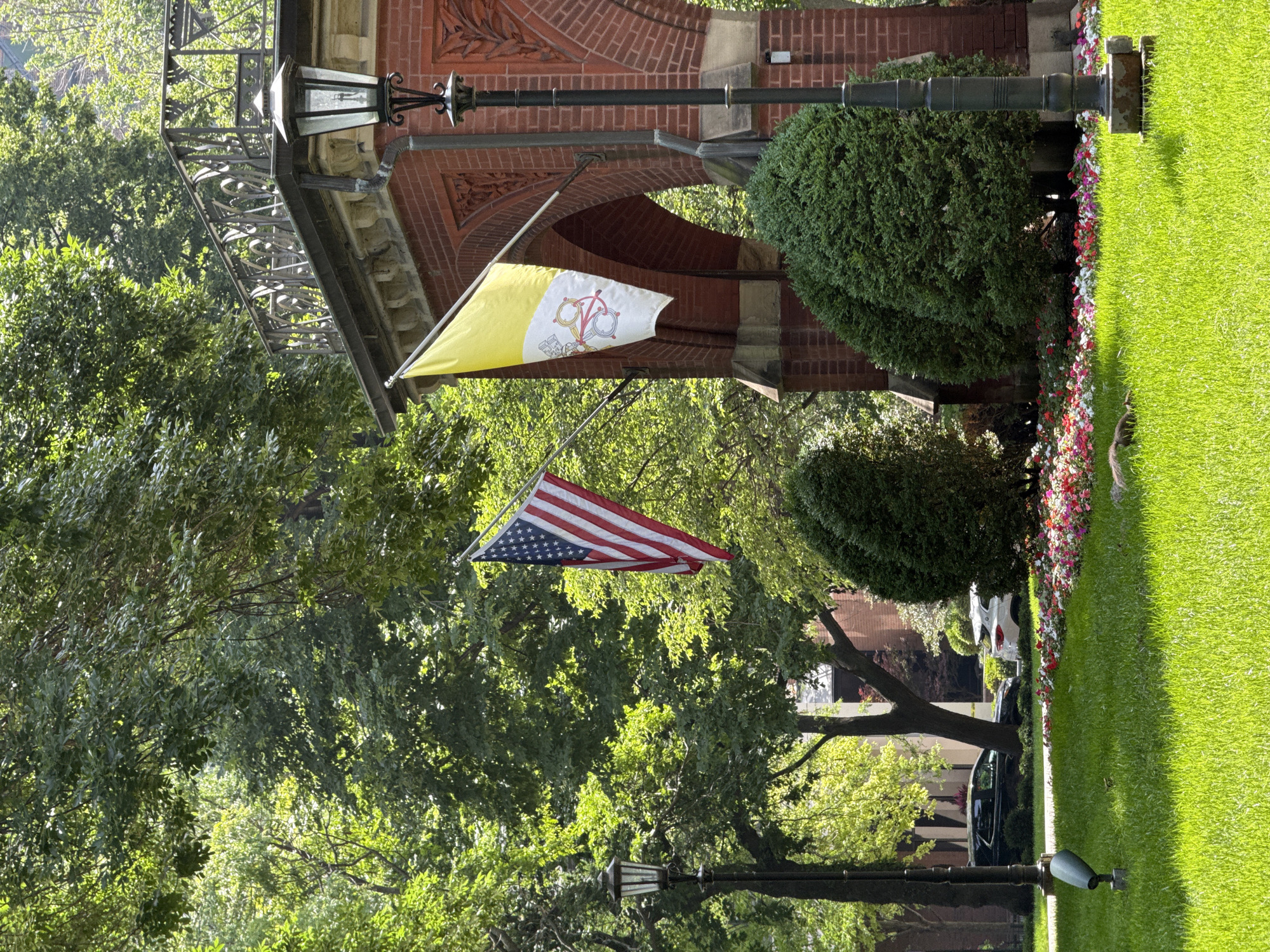 The image shows an exterior view of a building with a red brick facade, possibly a church or institution. Two flags are prominently displayed: one is the Vatican City flag, featuring a yellow and white bicolor with the Vatican's coat of arms, and the other is the flag of the United States of America. The flags are hanging from poles attached to the building.
The scene is set against a backdrop of lush green trees and manicured lawn, suggesting a pleasant day. The sunlight filtering through the leaves indicates it is daytime, likely late morning or early afternoon. A decorative wrought-iron balcony or railing is visible above the flags, and a lamppost is positioned in the foreground. Further back, a small flower bed adds a touch of color to the landscape. A dark sedan and a white car are partially visible in the background, suggesting the presence of a road or parking area. The overall impression is one of a well-maintained, possibly historic, property. There is no visible text in the image.