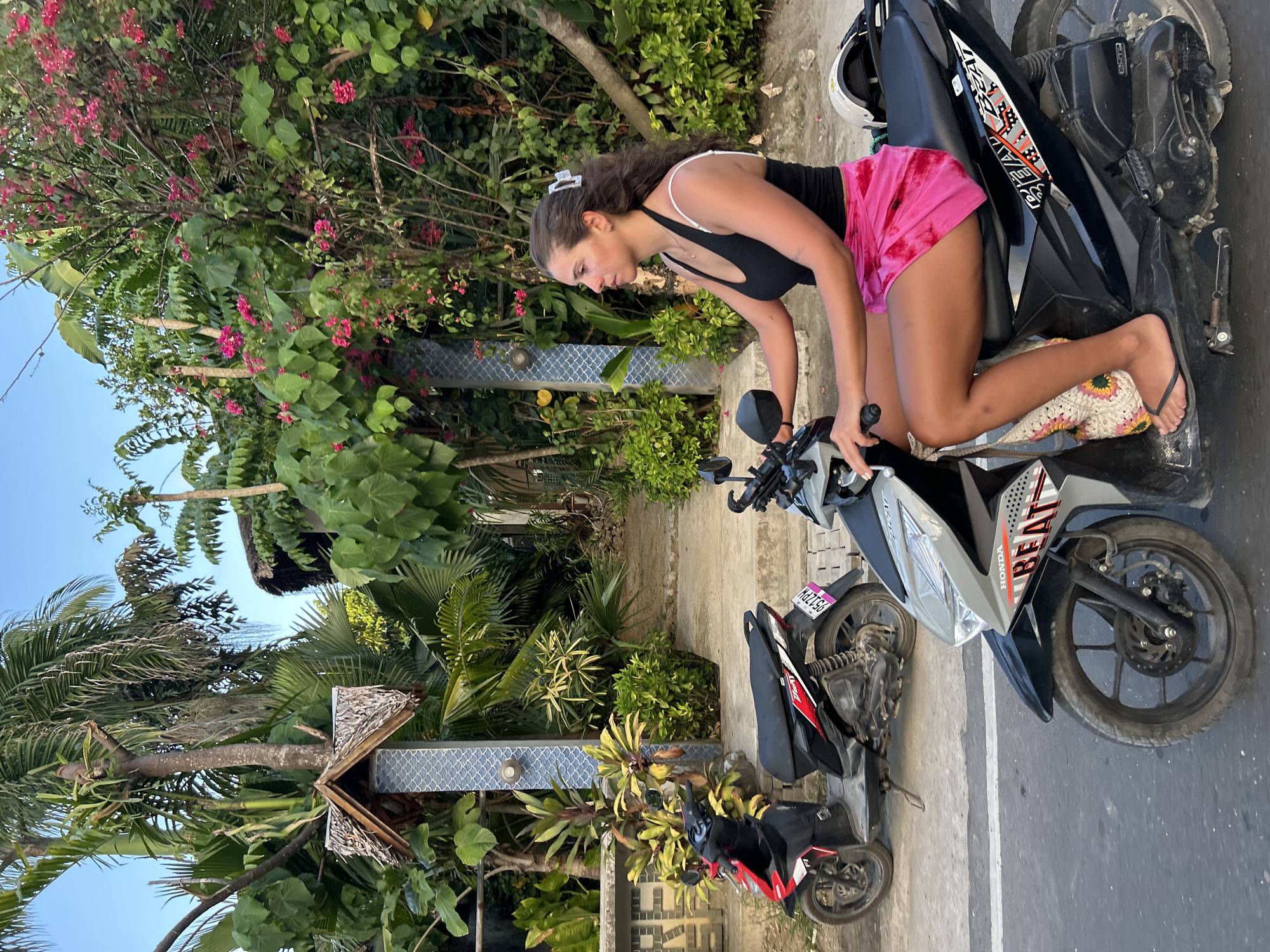 The image, originally rotated ninety degrees clockwise, captures a young woman on a scooter in General Luna, Philippines. The woman has dark hair, styled with a white claw clip, and is wearing a black tank top with white straps and pink tie-dye shorts. She is positioned on a silver and black Honda Beat scooter, leaning forward towards the handlebars, with flip-flops on her feet. A white helmet is placed on the rear seat of the scooter.
In the background, a second, red and black scooter is partially visible, displaying a pink license plate with the numbers and letters "0512PH". The scene is characterized by dense, tropical vegetation, including tall palm trees, various green bushes, and flowering plants with small pink blossoms. A wall with light blue patterned tiles and a rustic wooden structure with a thatched roof can be seen amidst the foliage. The ground appears to be a paved surface with a white line. The clear blue sky indicates a daytime setting, possibly late morning or afternoon. The overall atmosphere suggests a warm, vibrant outdoor environment.