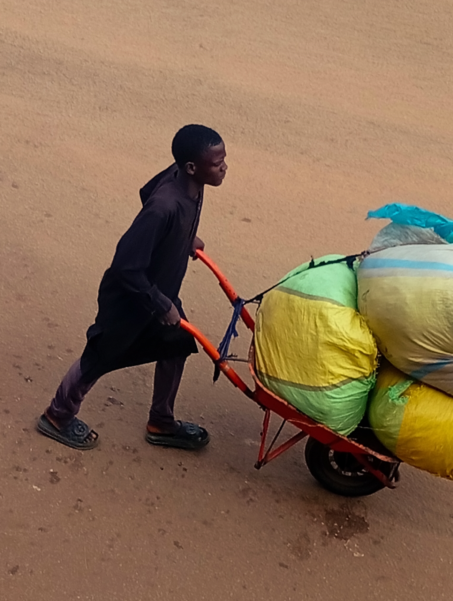 In Bukuru, Nigeria, a young person dressed in dark clothing and purple trousers is pushing a wheelbarrow laden with several large, colorful sacks. The wheelbarrow has bright orange handles and a black tire. The sacks are predominantly green, yellow, and white with blue and yellow stripes, and one is covered with a light blue plastic. The ground appears to be a dirt or asphalt road, with some scattered debris. The person is captured in profile, looking forward as they move the loaded wheelbarrow.