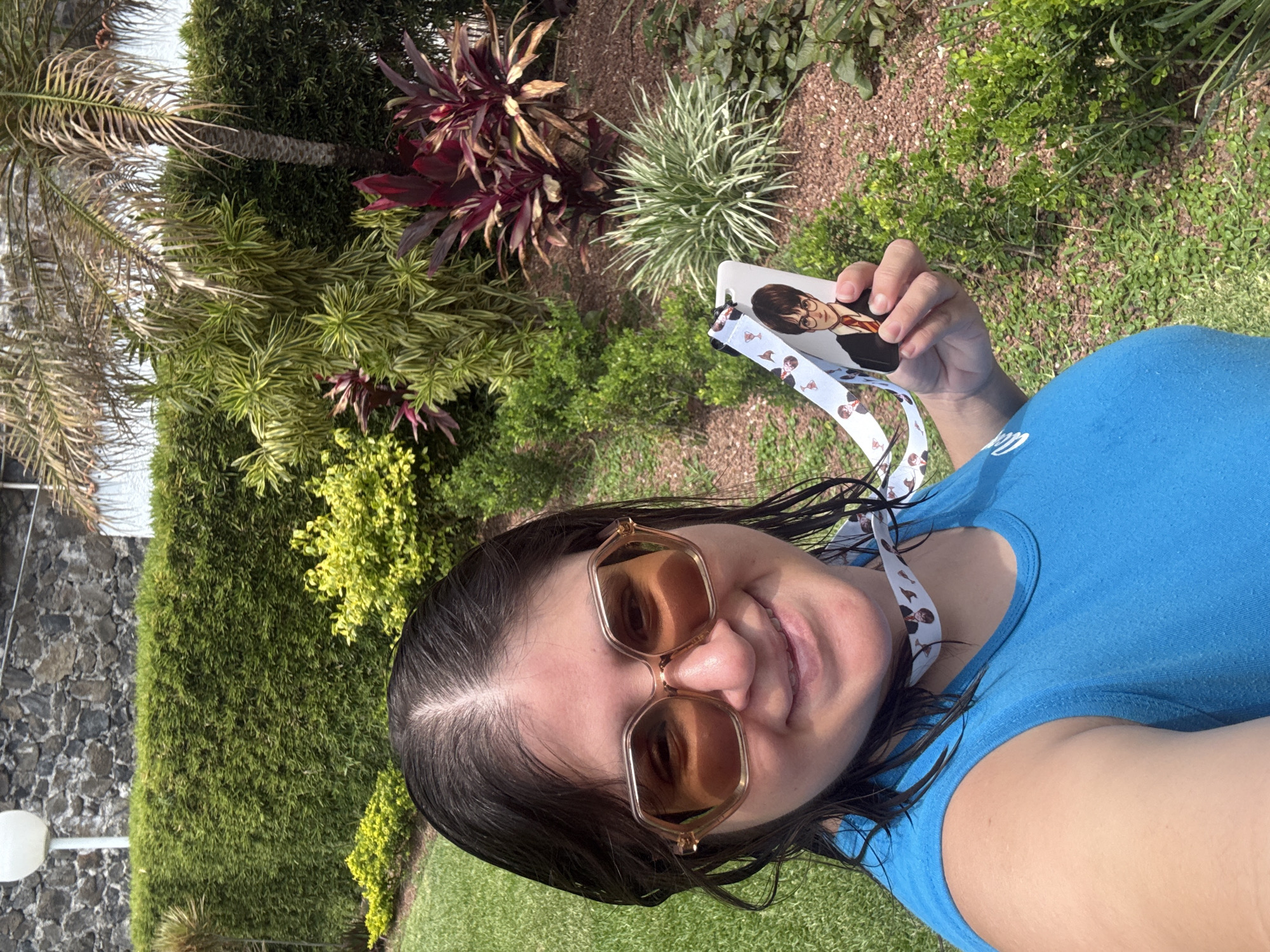 A woman is taking a selfie outdoors, holding up a lanyard and ID card with an image of Harry Potter. She is wearing sunglasses and a blue t-shirt. The background consists of lush green foliage, including bushes, grass, and various plants. A stone wall and a lamppost are also visible in the background. The location appears to be in a well-maintained garden or park. The city is Tres de Mayo, Mexico.