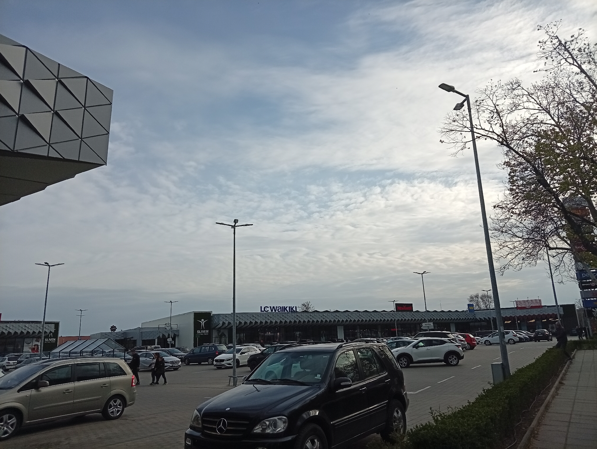The image depicts an outdoor scene at a shopping center in Sliven, Bulgaria. The sky is overcast with scattered clouds. Several cars are parked in the parking lot. In the center of the image, the LC Waikiki logo is visible above the building. A black Mercedes SUV is in the foreground. There are bare trees on the right side of the image. Streetlights are also visible. A section of a modern building with triangular paneling is on the left. People are walking in the parking lot. The ground is paved.