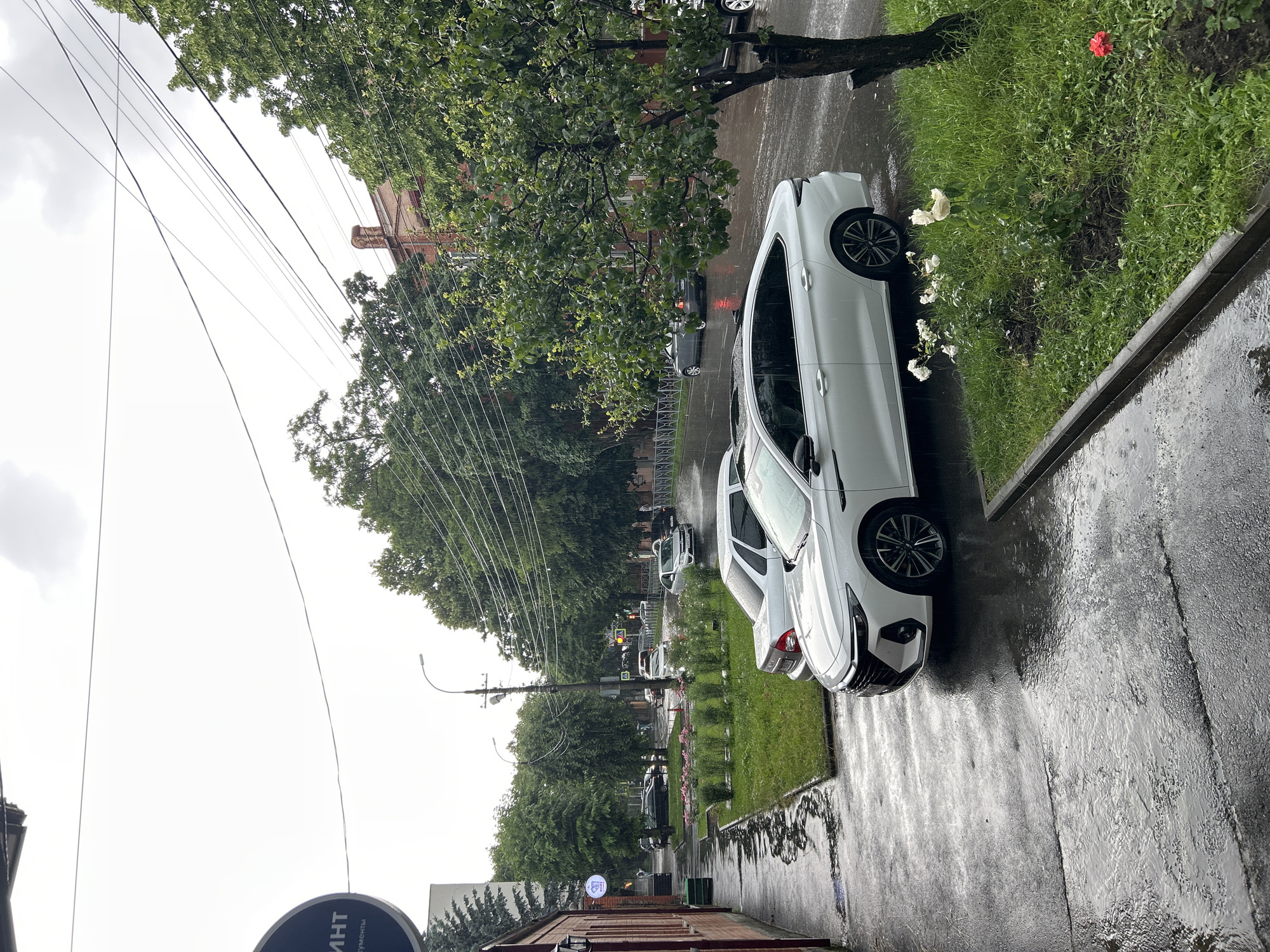 The image depicts a wet street scene in Oktyabrskoye, Russia, on an overcast day with light rain falling. Several white cars are parked along a sidewalk lined with green grass and trees. In the background, more cars are visible on the street, and a traffic light shows a red signal. A brick building is partially visible in the foreground. Overhead, power lines crisscross the sky. The overall mood is subdued, likely due to the gloomy weather. The scene appears to be an ordinary day in a residential or commercial area. The text "ИНТ" is visible on a circular sign hanging from a pole.