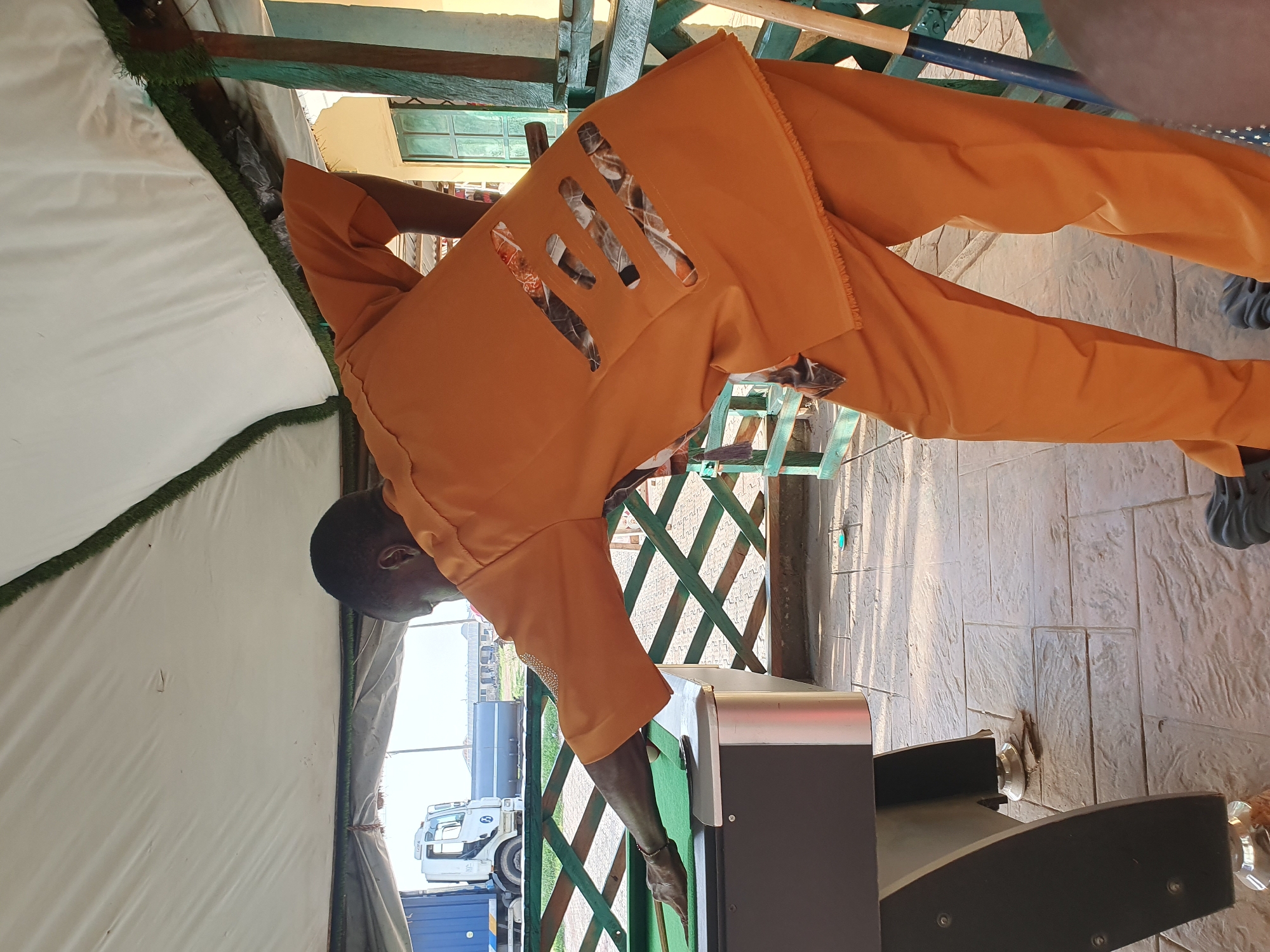 A Black man wearing an orange outfit is playing pool at an outdoor venue, possibly a bar or recreational area, with a light-colored tent providing overhead cover. The ground is paved with textured tiles. In the background, a white truck and a blue shipping container are visible. The man is positioned on the right side of the image, facing away from the camera, with his left hand on the pool table and his right arm extended towards a pool cue on the green felt. The outfit features a camouflage pattern visible through cutouts on the back. The overall lighting suggests it is daytime, with sunlight casting shadows on the ground. The image appears to be taken in Lokoja, Nigeria.