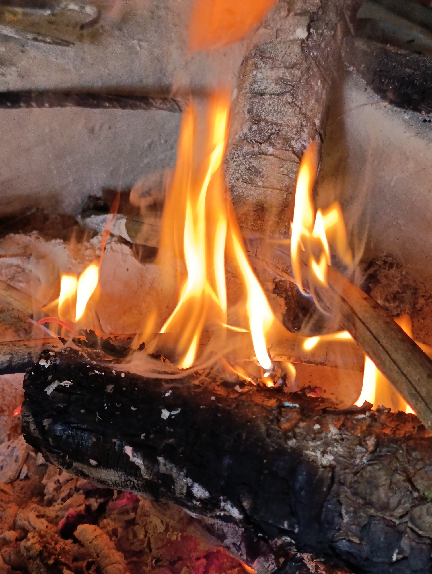 This is a close-up, medium shot of a fire burning in a fireplace. The flames are a vibrant orange and yellow, licking at charred logs. Ash and embers are visible in the foreground, with rough, textured stone forming the background. The image appears to be taken in Vashisht, India, a town known for its hot springs and temples.