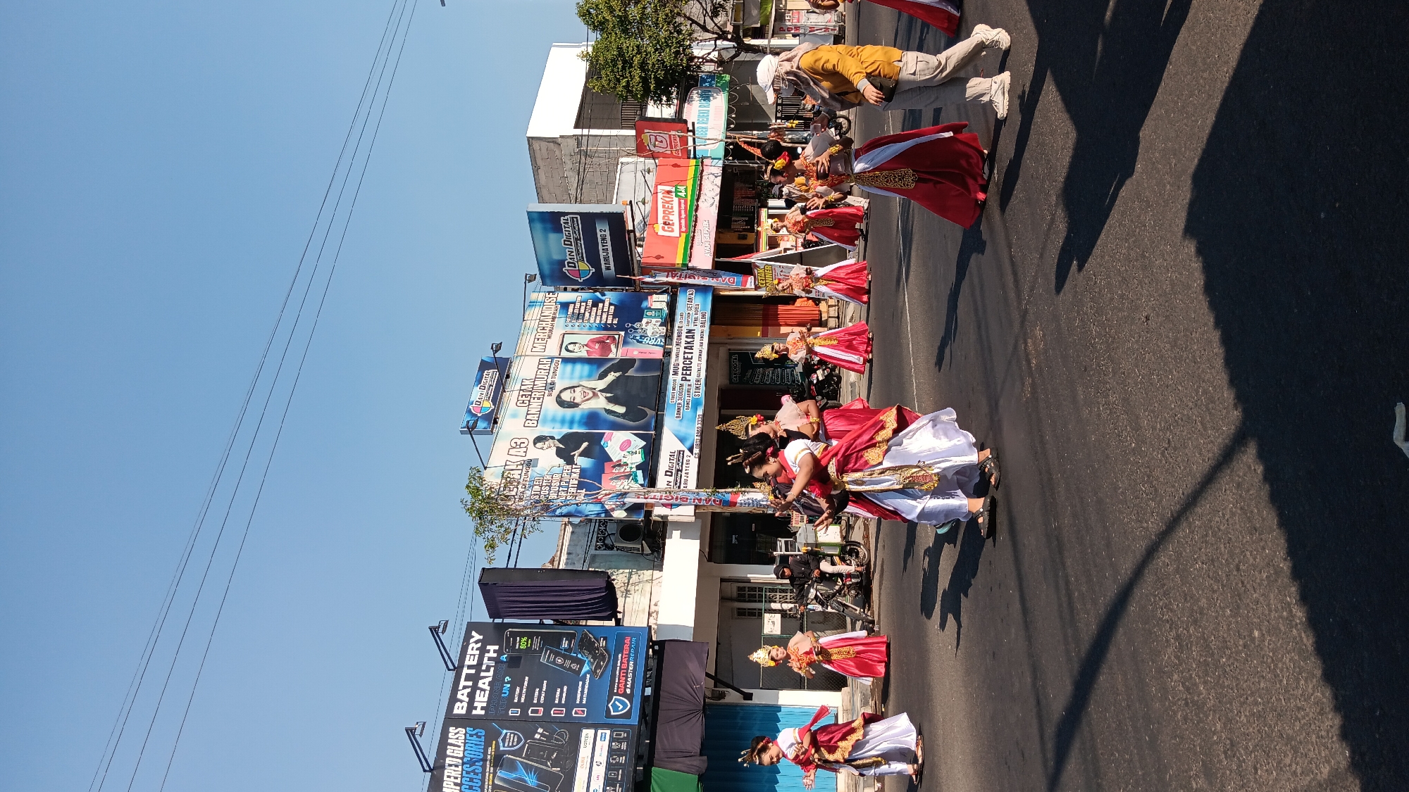 A group of dancers in traditional red and white attire are performing in a street procession in Warujayeng, Indonesia. The dancers, adorned with gold headpieces and intricate gold trim on their costumes, are captured mid-movement, with some holding small props. They are walking on an asphalt road in front of buildings displaying various advertisements. Notable signs include "DANI DIGITAL" banners advertising printing services, a sign for "BATTERY HEALTH" and "TEMPERED GLASS ACCESSORIES," and a "GEPREK" restaurant advertisement. A few motorcycles are parked along the side of the road, and a person in a yellow shirt and light-colored pants is walking on the right side of the frame, observing the procession. The scene is under a clear blue sky, with shadows cast on the road by overhead power lines and the buildings.