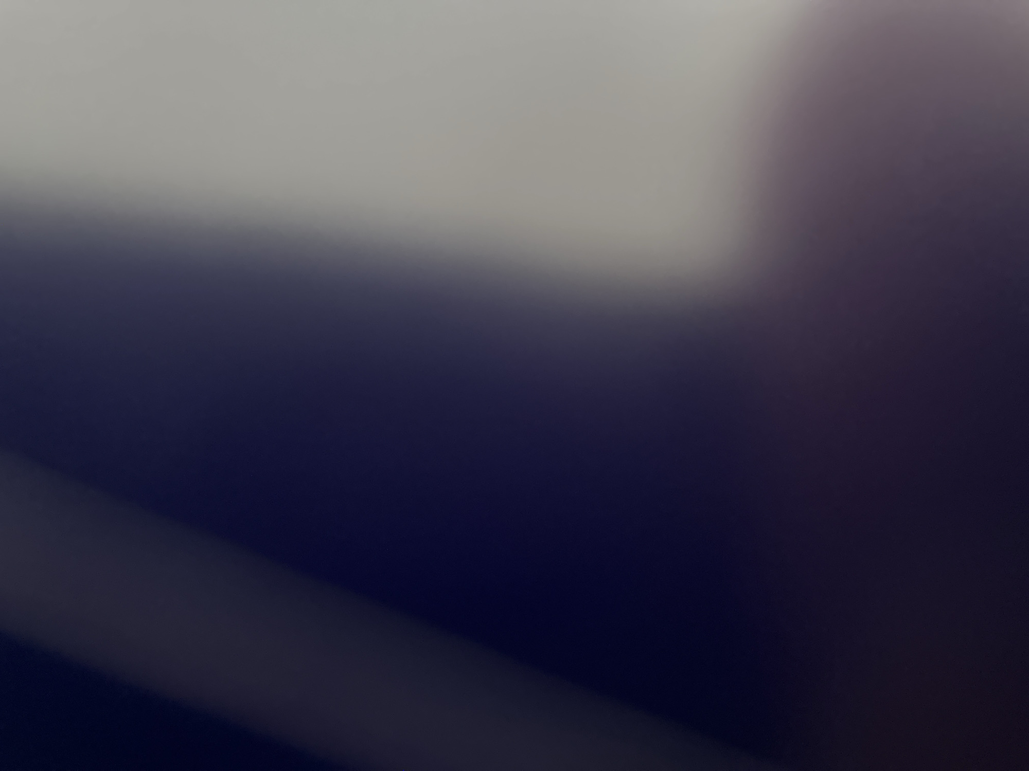 This is a close-up, abstract photograph with a gradient of color from a light gray at the top to a deep navy blue at the bottom. A diagonal shadow or light source creates a distinct band of lighter color across the dark blue area, starting from the bottom left and extending towards the upper right. The image is heavily blurred, with no discernible subjects, objects, scene, or setting. There are no visible text elements. The abstract nature of the image prevents any interpretation of activity, time of day, weather, location cues, or emotions.