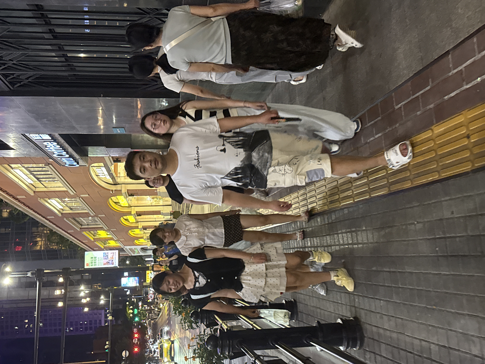 In Yuzhong District, China, at night, a group of people walks along a sidewalk adjacent to a street. The sidewalk is paved with dark grey planks and bordered by a raised tactile paving strip. On the left side of the sidewalk, a black metal railing separates it from a street with visible traffic lights and vehicles. In the background, buildings line the street, with illuminated signs and windows suggesting commercial establishments. The group consists of at least five individuals, including men and women of various ages. One man in the foreground wears a white t-shirt with a graphic print and patterned shorts. To his left, a young woman in a black and white dress walks alongside him. Further back, other individuals are seen walking, some carrying bags. The lighting is dim, with streetlights and building lights illuminating the scene.