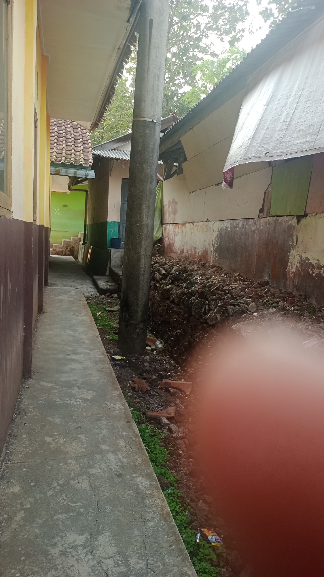 The image shows an exterior view of a narrow alleyway between two buildings. On the left, a building with a yellow facade and a brown lower section is visible. The alley itself is paved with concrete. On the right side of the alley, there is a large pile of debris, consisting of rocks, broken bricks, and other assorted refuse, next to a weathered, multi-colored wall. A tall, gray concrete utility pole stands in the middle of the alley, partially obscuring the view of the buildings behind it. The background reveals the corrugated metal roof of one building and the white tarp covering another. The overall impression is one of an unkempt outdoor area, possibly a service alley or backyard in a residential or commercial setting in Karyamandala, Indonesia.