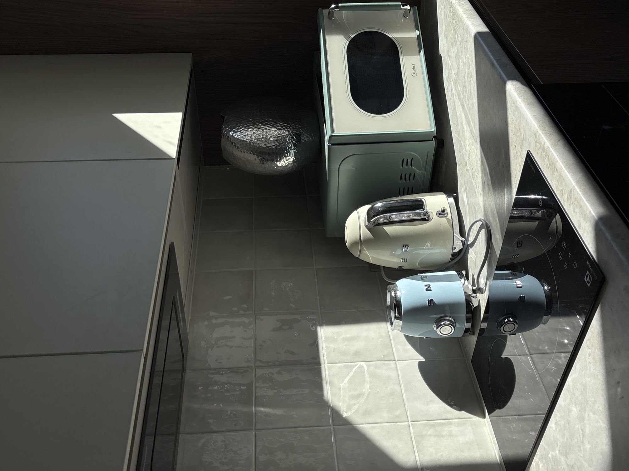 The image shows a kitchen setting with several appliances on a tiled floor. From left to right, there's a portion of a white cabinet with a visible corner. Next to it, there is a hammered metal bowl, a light-green Midea-branded microwave, a light-cream toaster, and a light-blue Smeg kettle. A portion of a black cooktop is visible on the right side of the image, with the Smeg kettle reflected on its surface. The scene is lit by bright sunlight creating distinct shadows. There is no information to identify the location of this image as the city of Krasnoyarsk, Russia.
