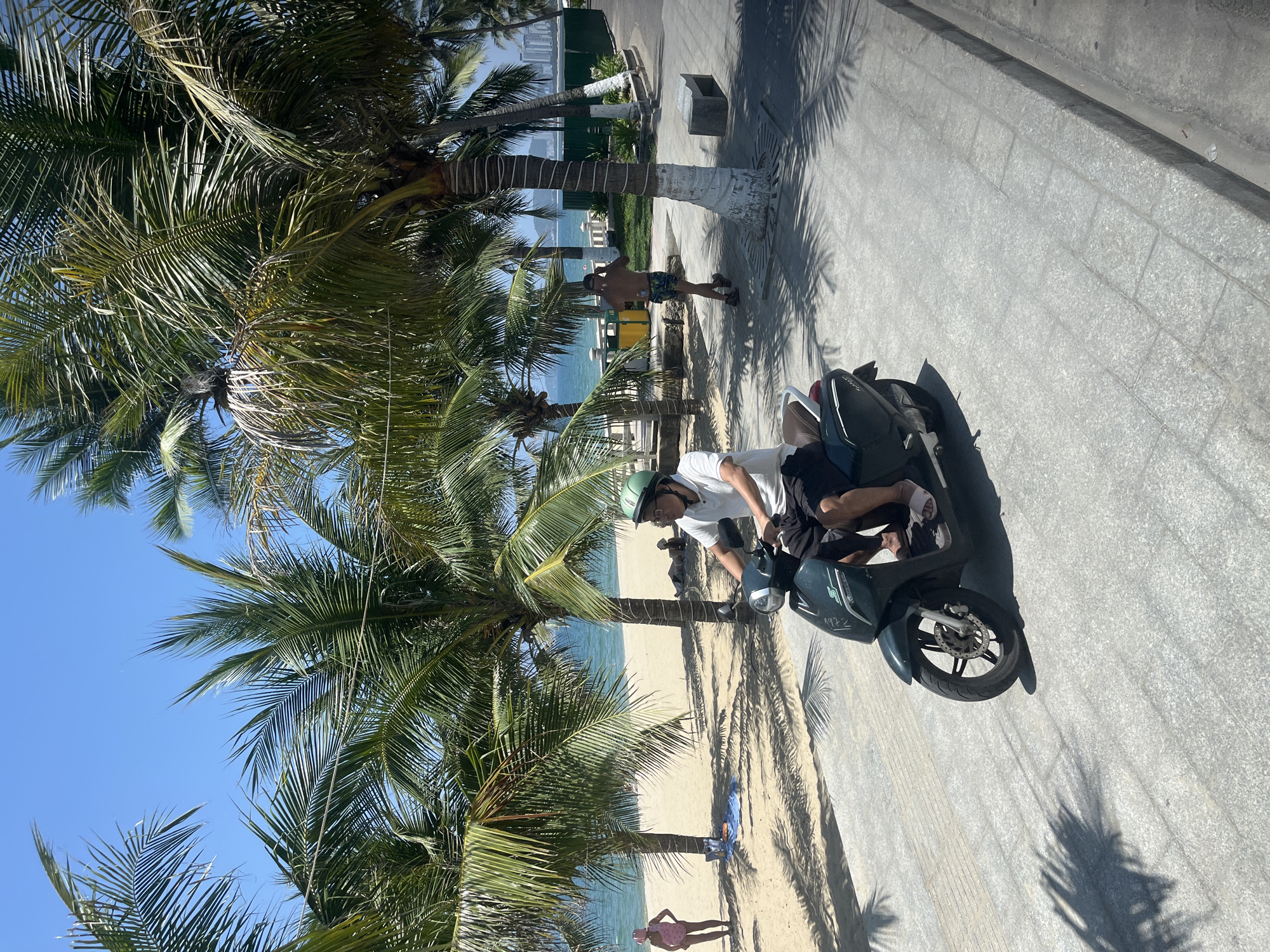 The image captures a sunny day along a coastal promenade in Nha Trang, Vietnam, likely near Tran Phu Beach. In the foreground, a man wearing a white polo shirt, black shorts, and a green helmet rides a dark green scooter. He has glasses on and is looking forward. To the left of the scooter, a sandy beach is visible with palm trees casting shadows. A woman in a pink swimsuit and hat stands on the beach, and another person is lying down on a towel. The sea is a calm blue, and in the background, there are buildings along the coastline. Further down the promenade, a shirtless man in patterned shorts is walking away from the camera. The promenade itself is paved with grey interlocking stones, and a concrete bench is visible to the right. Palm trees line the promenade, their fronds rustling in the light breeze.