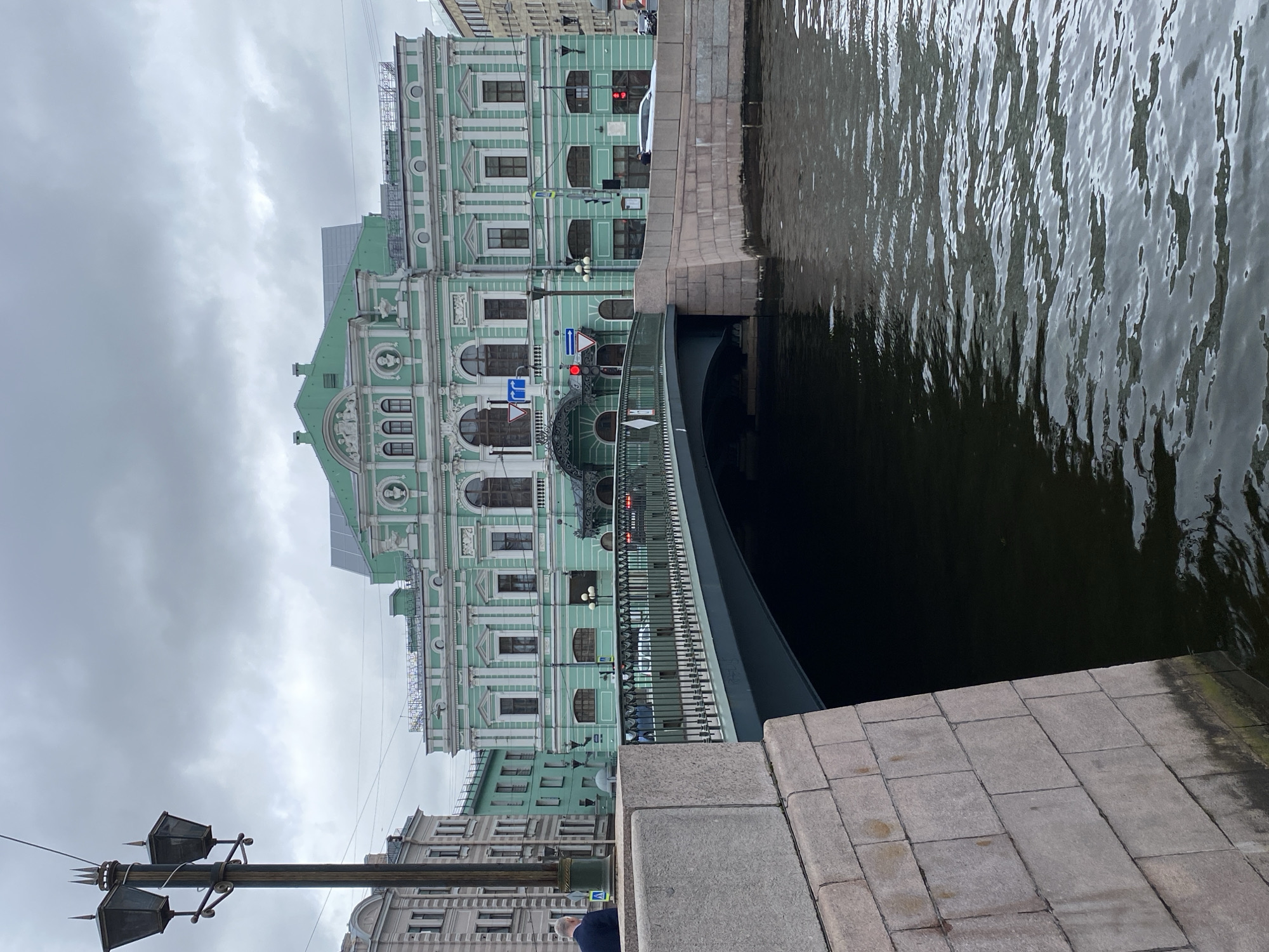 A wide-angle, eye-level exterior shot shows a building in Saint Petersburg, Russia, with a bridge and a canal in the foreground under a cloudy sky. The building, painted in a light green color with white decorative elements, features multiple stories with numerous windows, ornate architectural details, and a green roof. The bridge, made of grey metal and stone, arches over the dark, rippling water of the canal. A streetlamp with three lamps stands on the left side of the image, and traffic lights and signs are visible on the bridge. A few cars are driving across the bridge, and a person is standing on the left sidewalk, facing away from the camera. The canal's edge is lined with a stone embankment. The overall atmosphere is subdued due to the overcast weather.