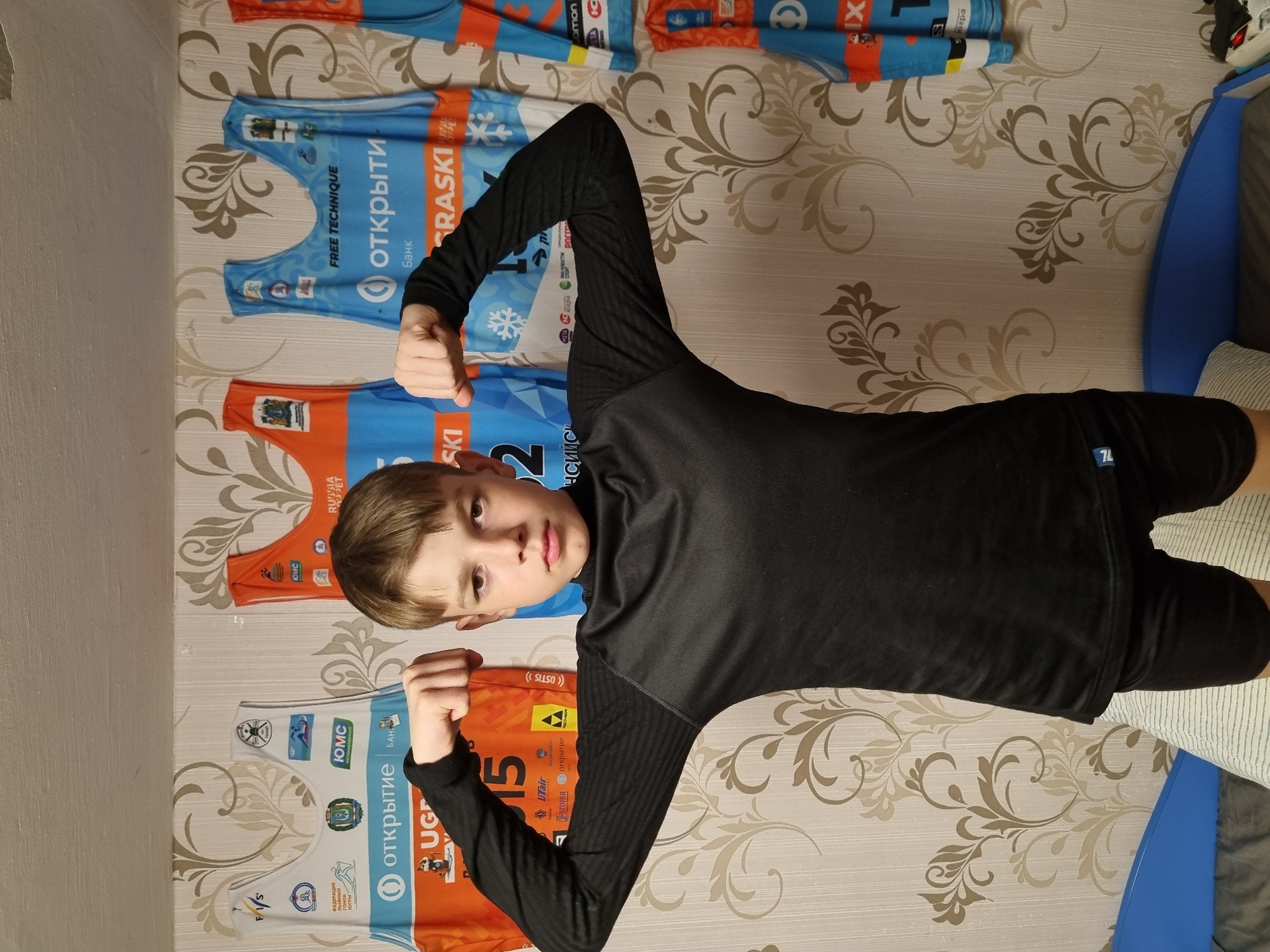 A boy is posing in front of a wall covered in patterned wallpaper. He is wearing a black long-sleeved shirt and black shorts. He is flexing both arms, creating a "muscle man" pose. Behind him, attached to the wall, are multiple athletic jerseys in shades of blue and orange, with text in Russian. According to the OCR data extracted from the photo, the location is in the city of Chaikovsky, Russia. The jerseys are likely related to cross-country skiing, as indicated by the text "лыжных гонок ЮГРЫ" and the presence of the term "SKI." Some of the visible logos include "ОТКРЫТИЕ," "RASKI," and "ЮГРЫ." A blue object (possibly a bed frame) is visible on the right side of the image.