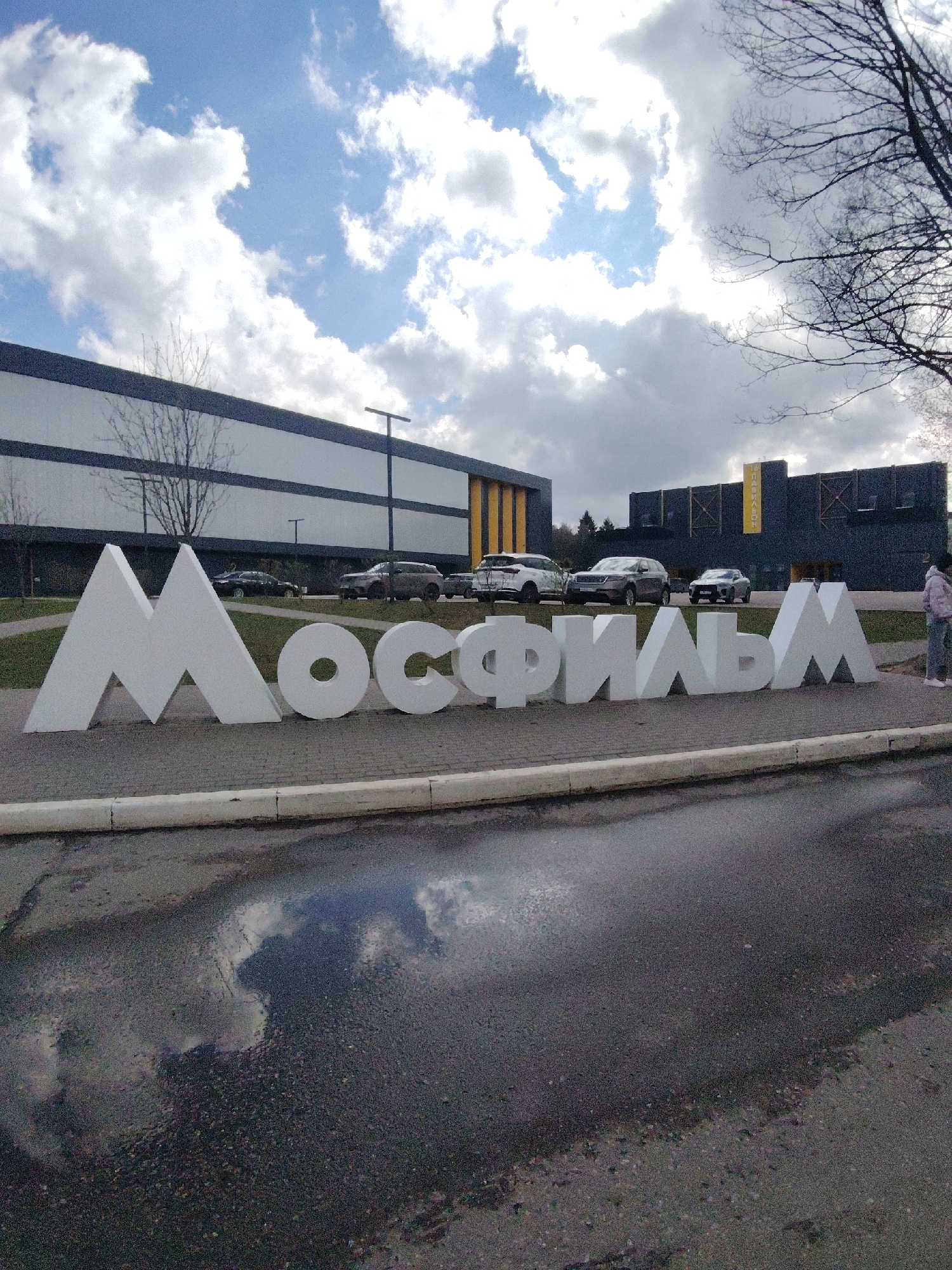 The image captures an outdoor scene featuring a large, white, volumetric sign spelling "МОСФИЛЬМ" in Cyrillic letters, placed on a grassy strip next to an asphalt path or road. This sign prominently identifies the location as the Mosfilm film studio in Moscow, Russia.
In the background, there are modern-looking, dark-colored buildings, possibly studio pavilions or administrative offices. One building on the left has horizontal reflective panels, while another further back and to the right is darker with some yellow accents and a yellow banner or sign partially visible, which appears to read "ПАВИЛЬОН" (Pavilion). Several cars, including SUVs and a sedan, are parked or driving in front of these buildings. The ground in the foreground is asphalt, showing wet patches and reflections, indicating recent rain or damp conditions. A curb separates the asphalt from the grassy area where the sign rests.
The sky above is partly cloudy, with patches of bright blue visible through fluffy white and light grey clouds. Bare trees with visible branches, indicative of early spring or late autumn, are scattered throughout the background, adding to the outdoor setting. On the far right, a person wearing a light-colored jacket and dark pants is standing near the end of the "МОСФИЛЬМ" sign, looking towards the left, providing a sense of scale. The overall impression is a clear daytime scene.