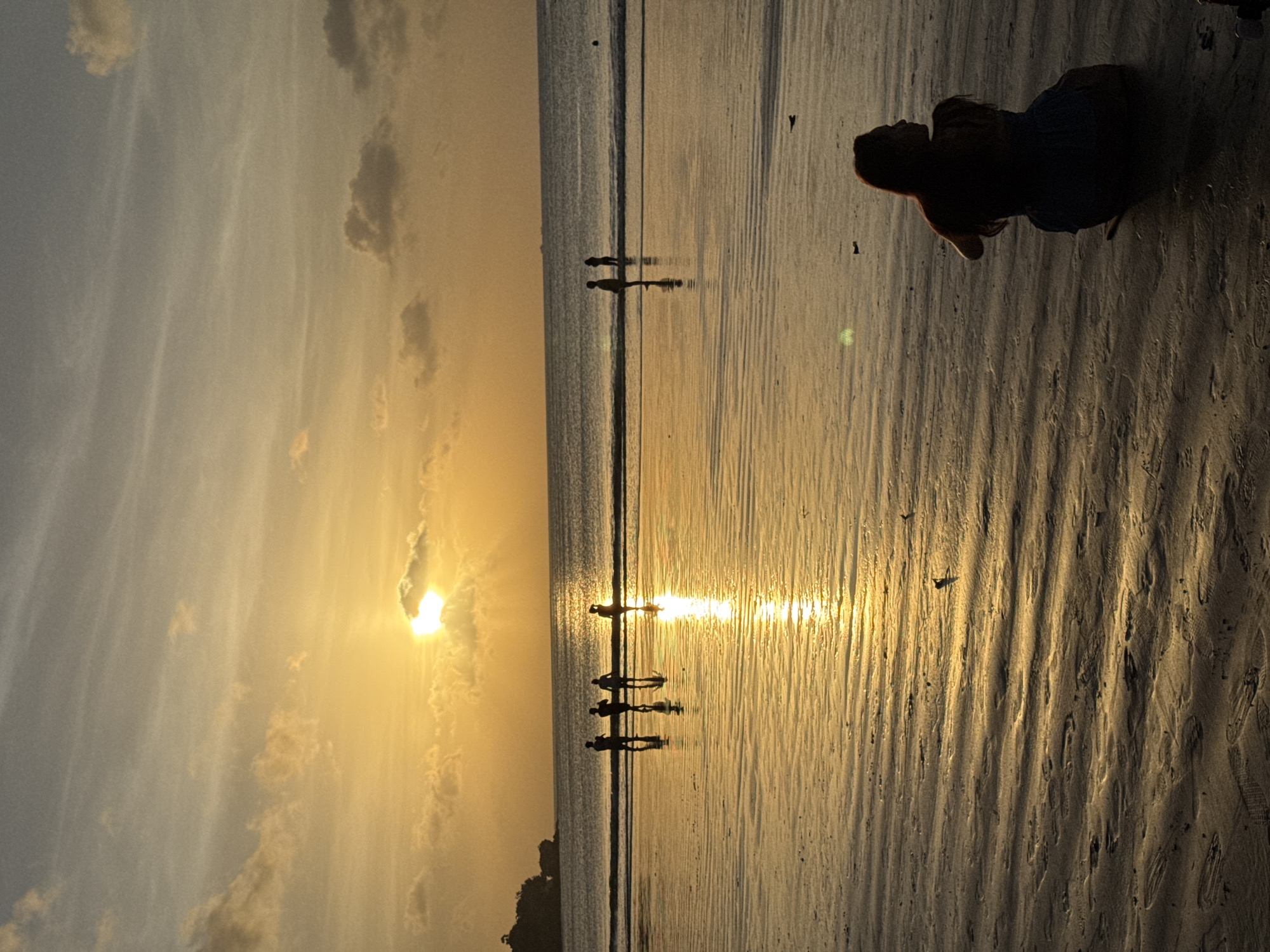At Kuta Selatan, Indonesia, a person is captured in silhouette, positioned in the lower right of the frame on a beach. The golden glow of the sunset reflects off the wet sand and water. Silhouettes of several individuals stand together in the water, further into the scene, as well as additional people further into the background. There are some clouds in the sky above the sunset.