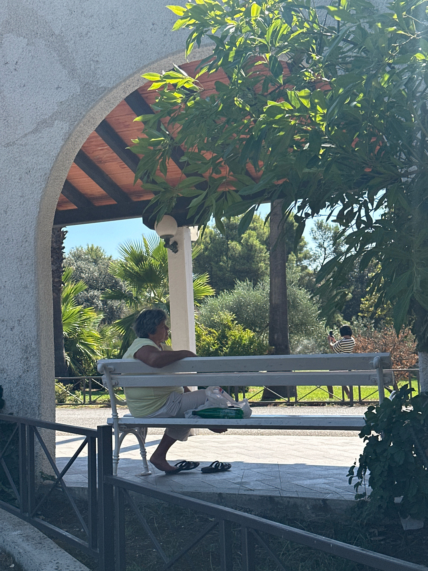 The image captures a serene outdoor scene, seemingly in Budva, Montenegro, under a clear blue sky. In the foreground, a white wrought-iron bench sits on a paved area, partially shaded by an arched structure with a wooden roof and a concrete wall. A woman with short, gray hair is seated on the bench, facing right, wearing a light yellow top and white trousers. Her right foot, clad in a black flip-flop, rests on the ground, while her left foot is bare. Next to her on the bench, a white plastic bag and a green object are placed.
Further in the background, another person wearing a horizontally striped shirt is visible, holding a dark rectangular object, possibly a phone, and appears to be taking a picture. The surrounding environment is lush and green, with palm trees, olive trees, and other dense foliage creating a natural backdrop. A dark metal fence runs along the lower portion of the image, separating the paved area from the grassy ground. Sunlight filters through the leaves of the trees, casting dappled shadows on the scene.