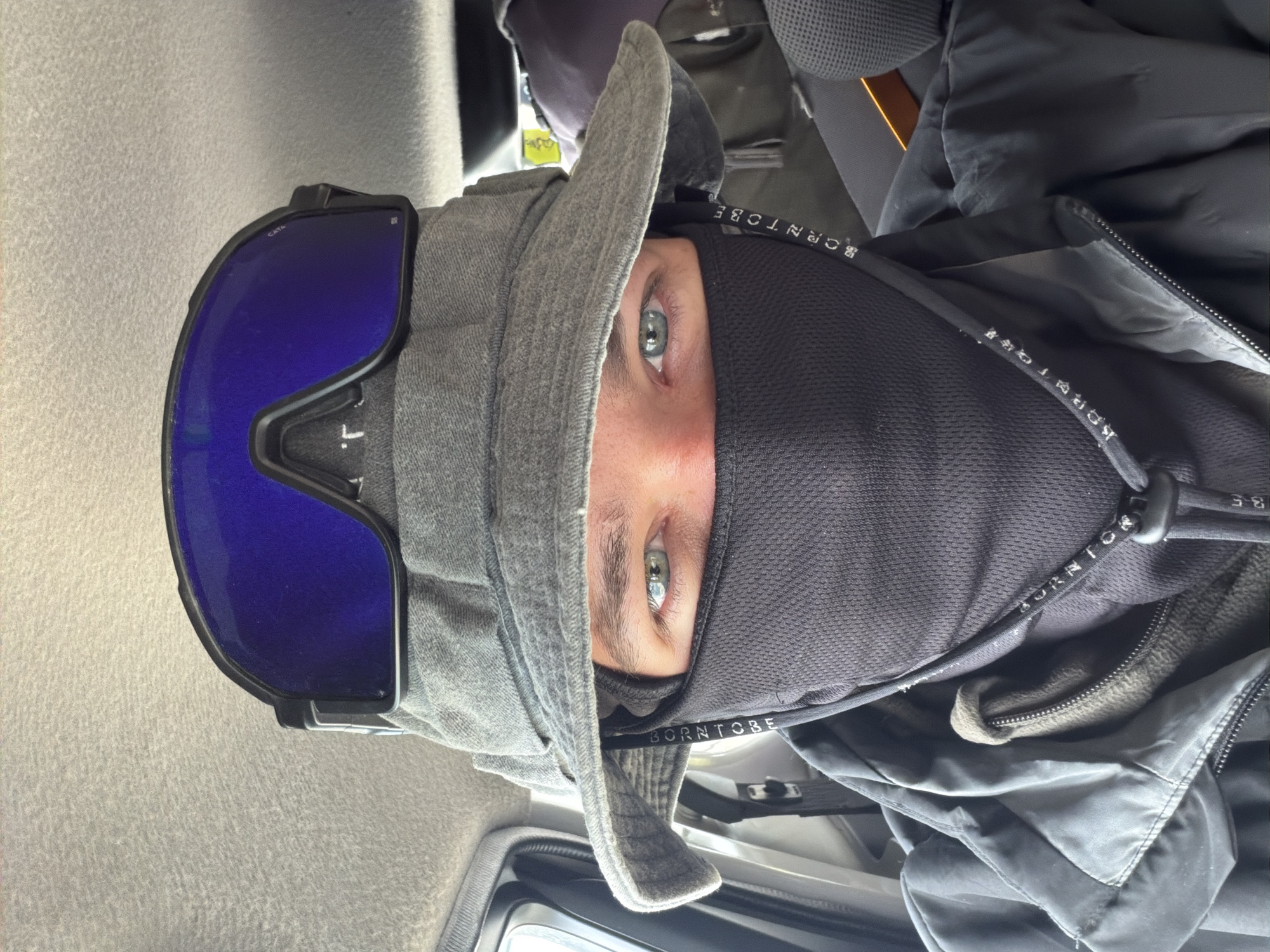 A man, partially obscured by a black face mask, grey bucket hat, and blue-tinted goggles, looks directly at the camera from inside a vehicle. The interior of the vehicle is dimly lit. The man is wearing a dark jacket, layered over a lighter olive-colored garment. The word "BORNT OBE" is visible printed vertically on a strap hanging from the mask. The background shows the grey interior of the vehicle, including seats and luggage. The overall scene suggests the man is either preparing for or has just concluded an outdoor activity, possibly in a cold or dusty environment. The location of Karzok, India is not determinable from the image itself.