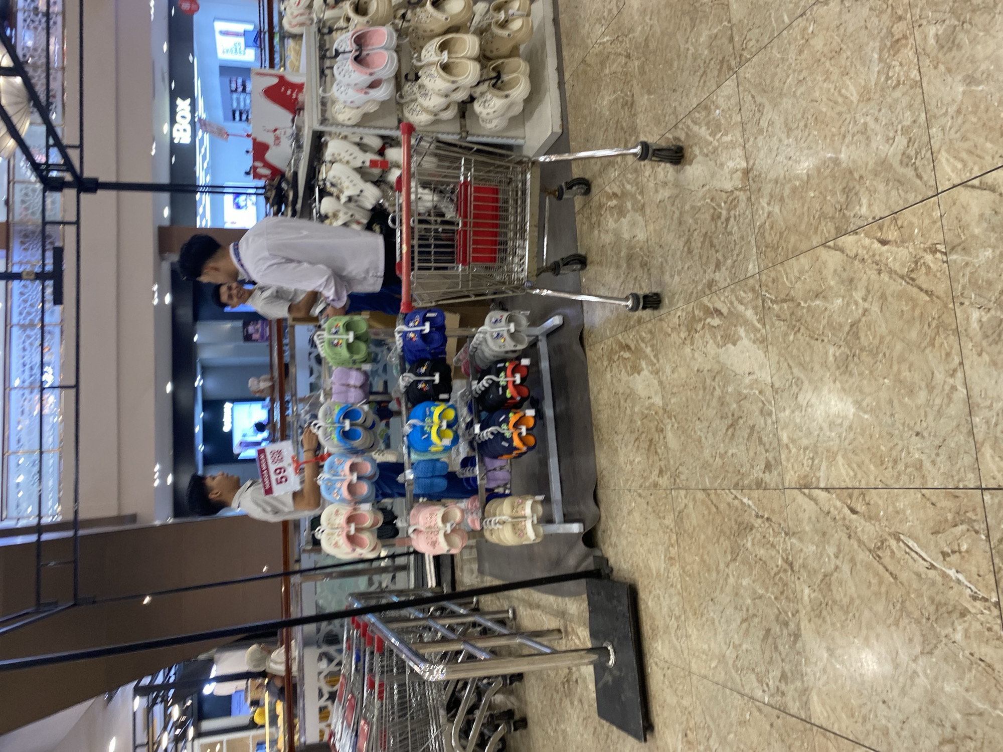 The image shows an indoor market stall in Banda Aceh, Indonesia, selling footwear. Two male employees, dressed in uniform shirts, are visible. One employee is interacting with a customer, while the other is looking away. The stall is stocked with various types of sandals and flip-flops, displayed on racks. A shopping cart is positioned in the foreground. The flooring is tiled with a marble pattern. The overall lighting suggests an indoor commercial setting, possibly a department store or a large market.