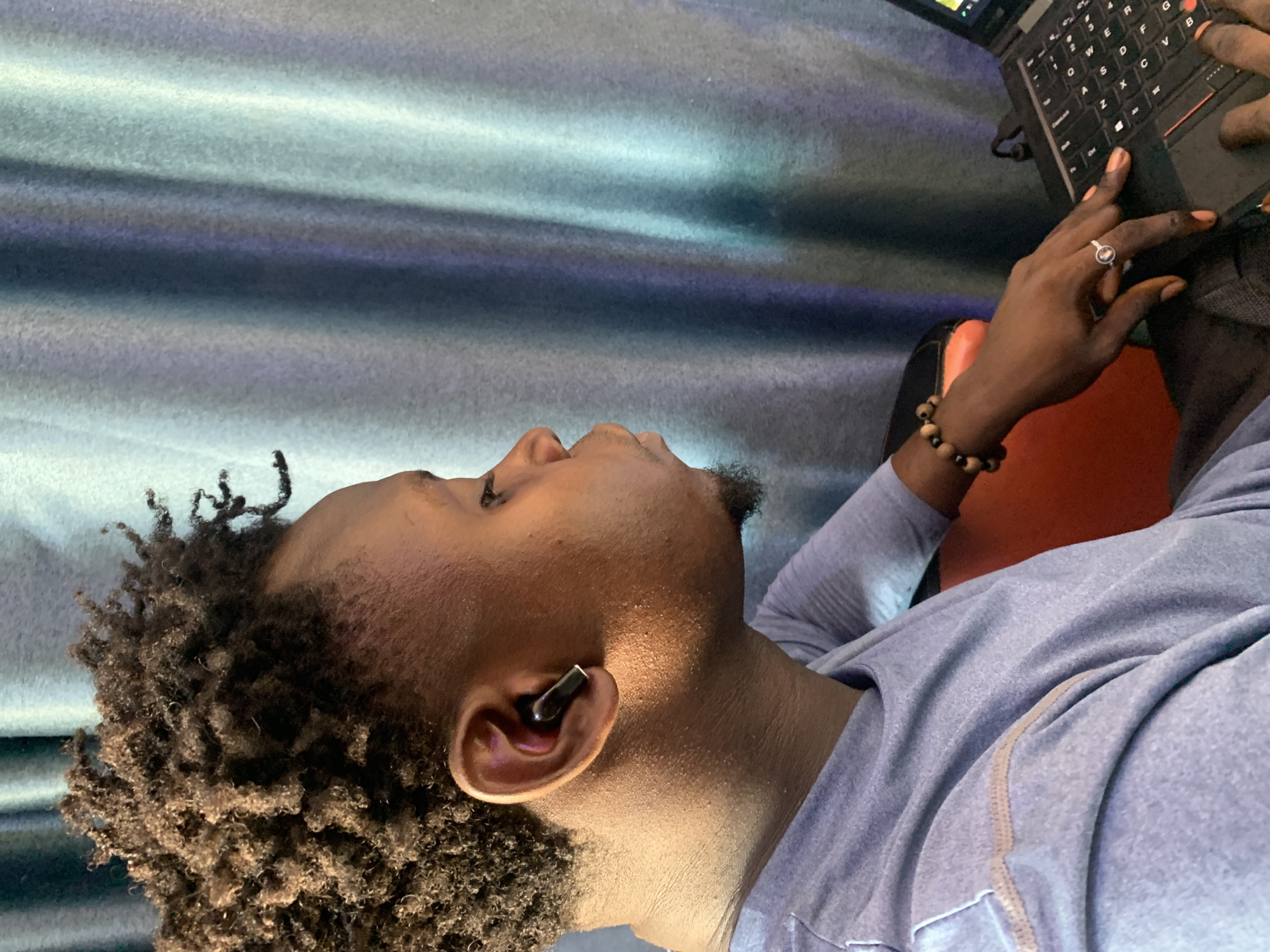 A person with dark skin and curly hair, wearing a light blue shirt, is using a laptop. The person is wearing an earbud in their left ear, a ring on their finger, and a beaded bracelet on their wrist. The background appears to be a wall or curtain with a blue and gray pattern. There is no indication that this photo was taken in Sabojibi, Nigeria.
