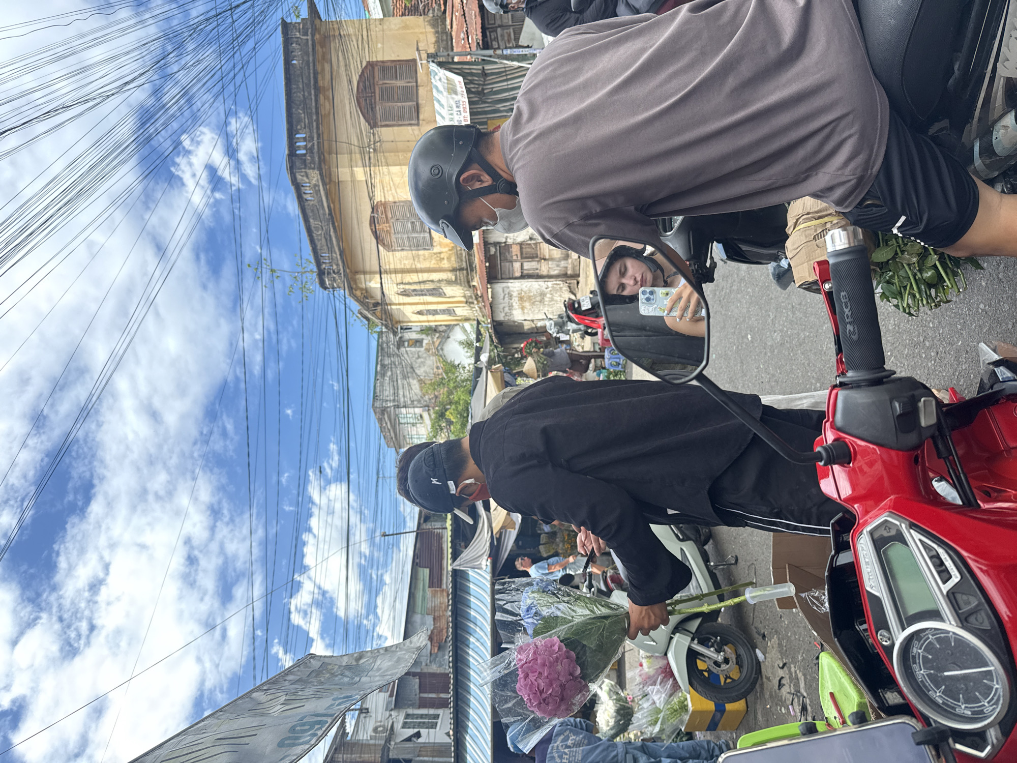 This is a street scene in Nha Trang, Vietnam, likely a market due to the presence of flower stalls and multiple individuals on motorcycles. The foreground features the handlebars and dashboard of a red motorcycle. A young woman with a helmet on is reflected in the motorcycle's mirror, holding a phone and taking a selfie. She is wearing a white tank top. To her right, a man wearing a grey t-shirt and black shorts, also with a helmet, is seated on the same motorcycle. In the mid-ground, two men are interacting. One, wearing a black shirt and a black cap with a red face mask, is holding a bouquet of pink and blue flowers. The other man is partially obscured. Several motorcycles, some with flower arrangements, are visible, along with market stalls displaying various flowers. The background shows buildings with aging facades and a sky with scattered clouds and overhead wires. The overall atmosphere suggests a bustling local market environment.