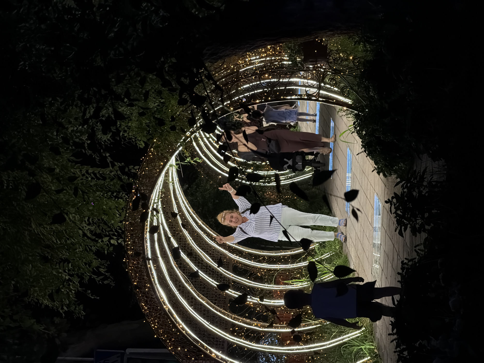 The image depicts an outdoor scene at night, likely in a park or public area, with a prominent illuminated archway decorated with string lights. A young boy is standing with his back to the camera, facing towards the archway. Further within the archway, a woman with blonde hair, wearing a striped shirt and light-colored pants, is facing the camera with her arms raised in a celebratory pose. Several other individuals, including two women and a child, are visible walking through the illuminated archway in the background. The ground appears to be paved with brick-like materials, and surrounding vegetation is dark and indistinct due to the nighttime setting. Some dark, leaf-like shapes are digitally superimposed throughout the image, obscuring portions of the scene. The overall atmosphere is festive, suggesting an evening event or attraction. This scene is located in the city of Chirchik, Uzbekistan, within the "Magic City" entertainment park.