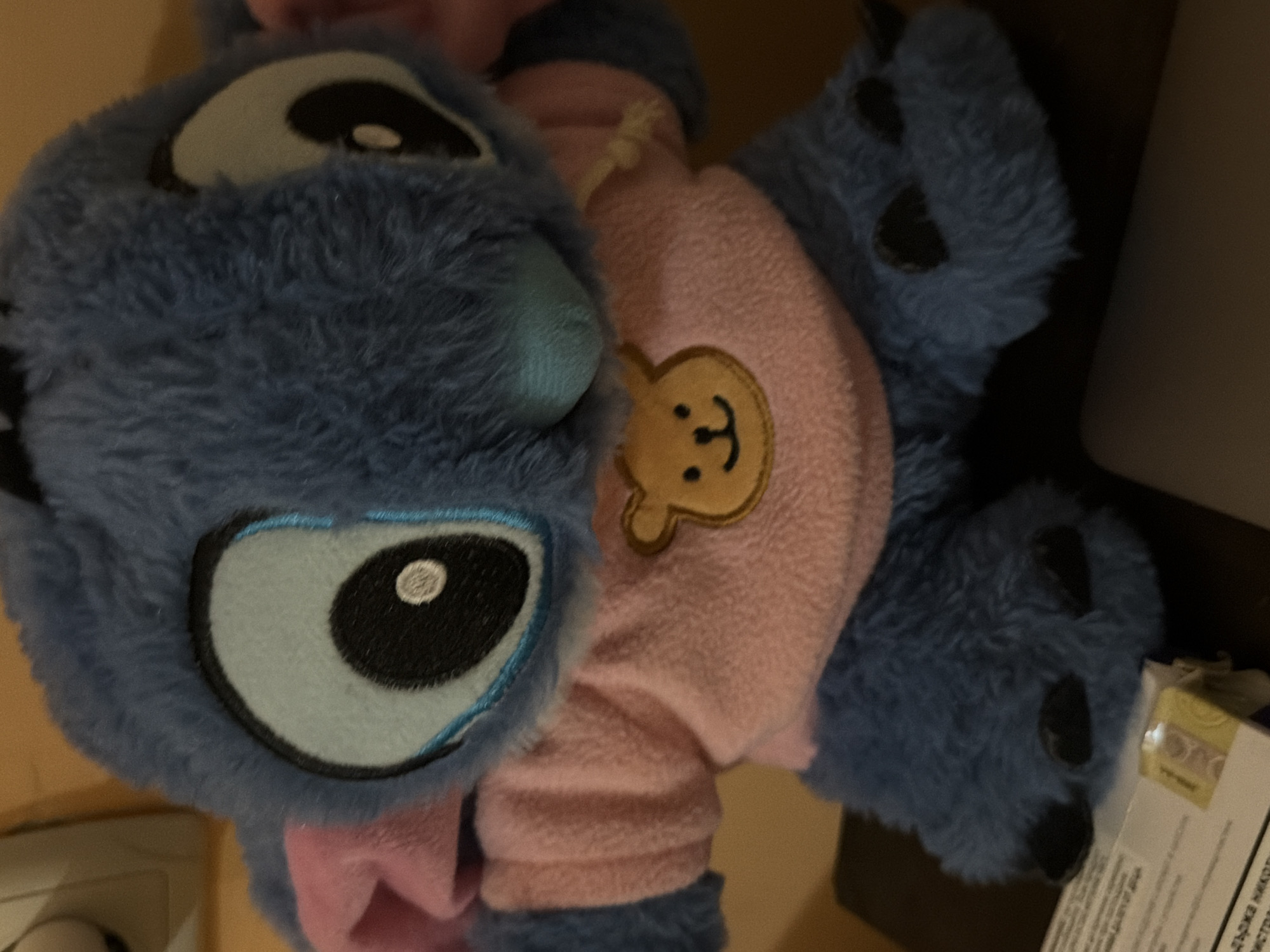 A close-up shot shows a blue plush toy, possibly Stitch from Disney's Lilo & Stitch, wearing a pink fuzzy outfit. The toy is positioned on its side, with its large, round eye clearly visible. The eye is black with a white pupil and is outlined in black and light blue stitching. A light blue nose is also visible. The pink outfit has a small, embroidered teddy bear face on the chest. The toy's fuzzy blue limbs are also visible, with black paw-like markings on the feet. The background is blurred, but appears to be an indoor setting, with a dark surface and what looks like a white box with some text on it in the lower right corner. The text on the box is partially visible and appears to be in Cyrillic script. The lighting suggests it is daytime or an indoor environment with artificial lighting. The overall impression is of a child's toy, suggesting a playful or domestic scene.