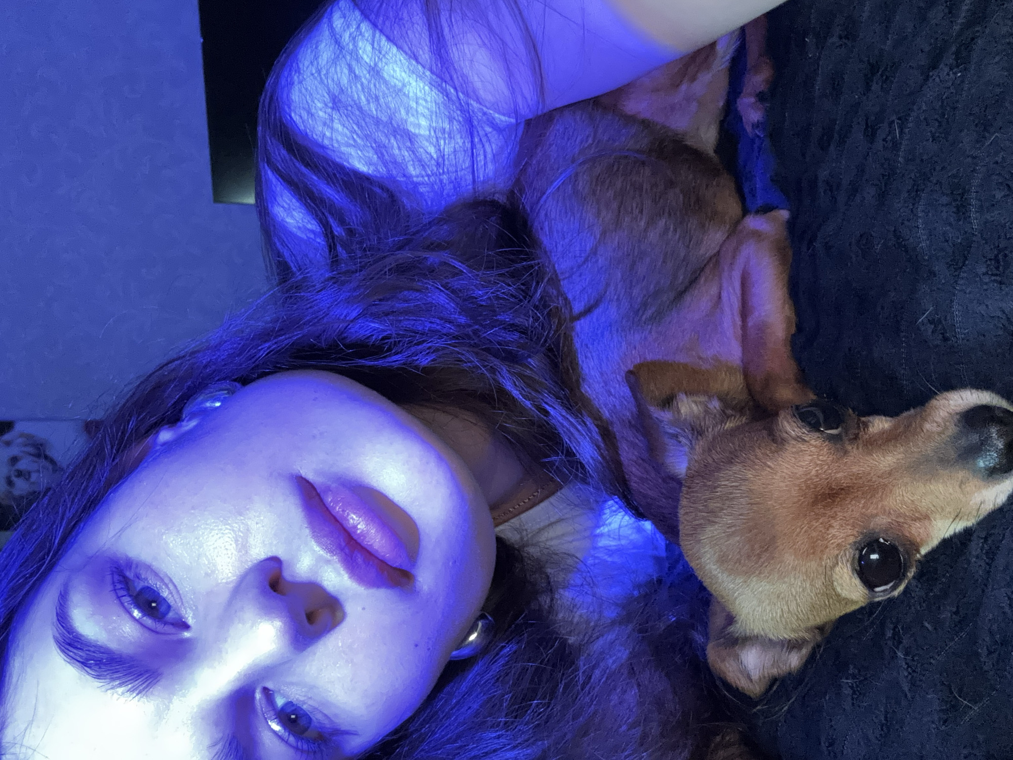 A close-up, low-angle shot shows a person and a dog lying down. The person, likely female, is positioned on the left side of the image, facing the camera with her head tilted. She has long, dark hair, light skin, and her eyes are open. The dog, a small breed with short brown fur, is on the right side of the image, with its head near the person's hair. The dog's head is tilted slightly upward, and its eyes are visible. The background is dark and indistinct, appearing to be a textured fabric, possibly part of a blanket or piece of furniture. The overall lighting in the image is blue, with a strong blue cast on the subjects. The location is unspecified and unknown.