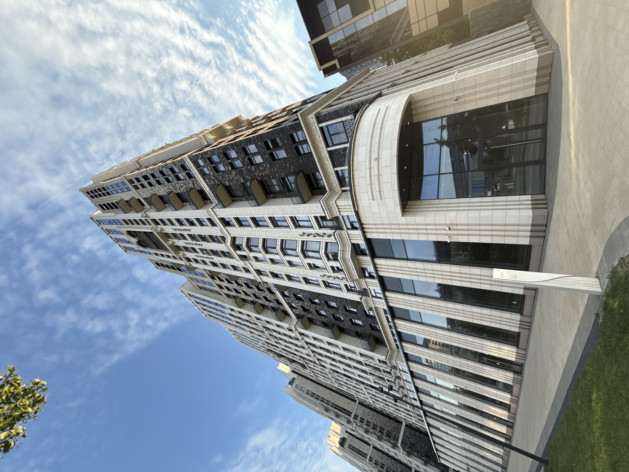 An extreme low-angle, full outdoor shot shows the exterior of a modern, multi-story residential building in Moscow, Russia, under a bright blue sky with scattered white clouds and the upper branches of a green tree. The building is tall and sleek, with a facade of alternating dark gray and light beige panels. It features numerous windows, many with black frames, and several balconies with dark railings. The lower portion of the building has large, floor-to-ceiling windows and a curved entrance area with a beige stone facade. A concrete sidewalk and a strip of green grass are visible in the foreground. There are no people or animals in the image. The scene appears to be a sunny day in a contemporary urban setting.