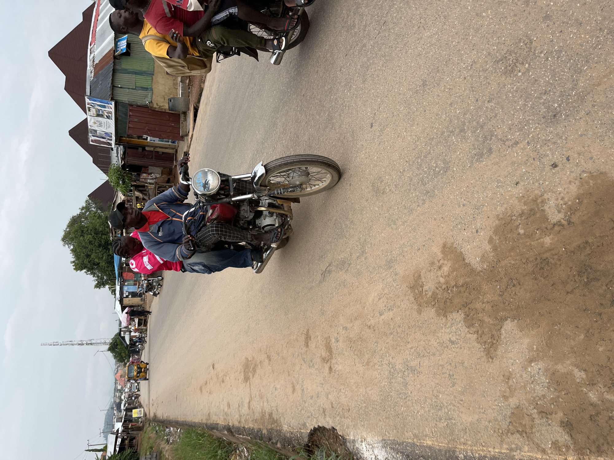 The image shows a street scene in Abuja, Nigeria. In the foreground, two motorcycles, each carrying two or more people, are traveling on a paved road. The road is worn, with patches and discolorations. The man riding the motorcycle in the center is wearing a navy jacket, jeans, and a dark cap. Behind him, another person wears a pink jacket. In the background, there's a street lined with shops and buildings, some with corrugated metal roofing. A tall communications tower is visible in the distance. The sky is overcast. The overall scene conveys a sense of everyday life in an urban area in Nigeria.