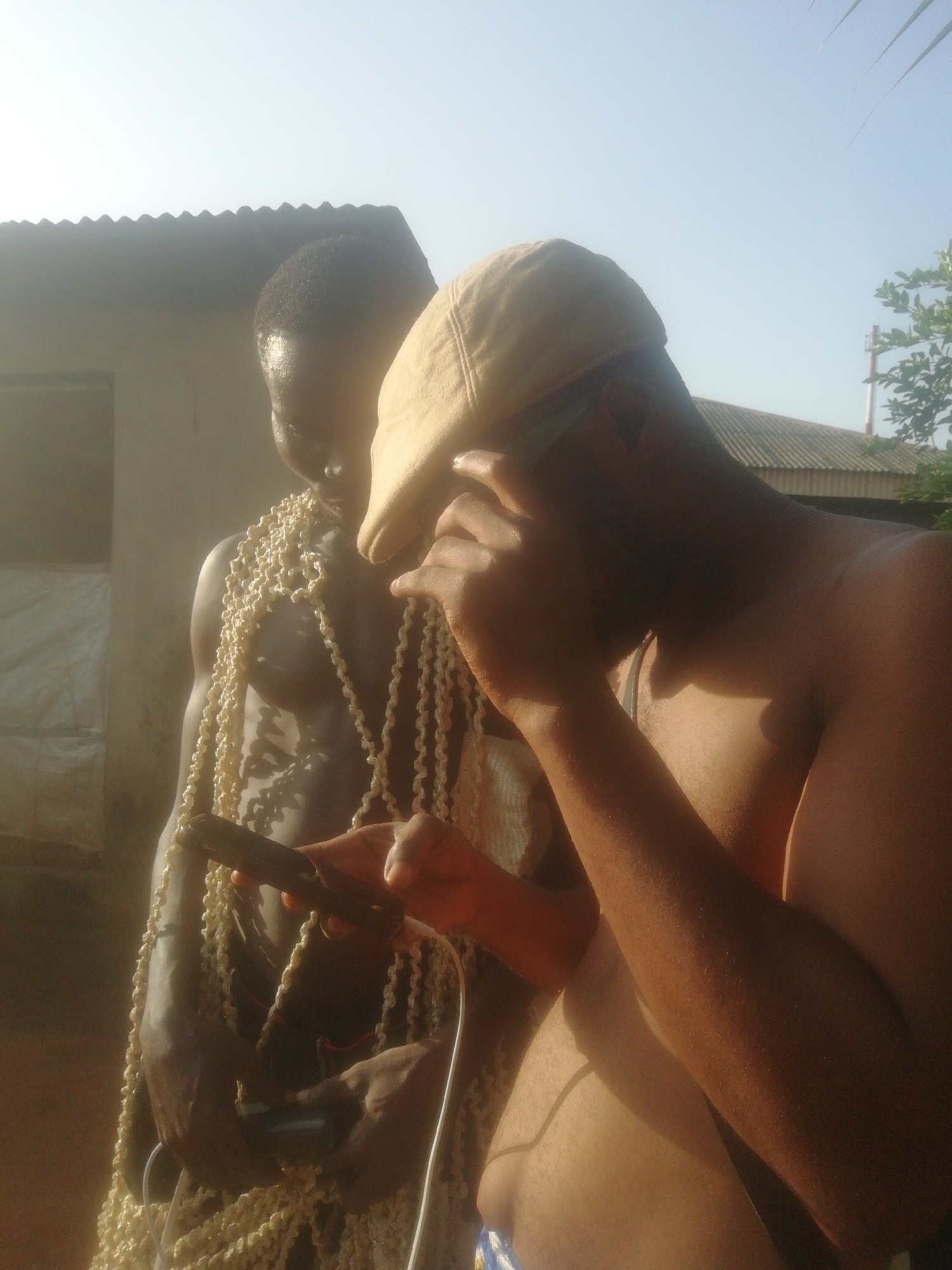 The photograph shows two shirtless men in an outdoor setting. The man in the foreground wears a tan baseball cap, obscuring part of his face. He holds what appears to be a homemade contraption consisting of wood, wires, and a small box in his hands. The second man stands behind him, partially obscured, and is adorned with multiple strands of what looks like small, off-white beads or similar material. The background features a building with a corrugated metal roof, suggesting a simple or rural structure. The lighting indicates a bright, sunny day. The location in the city of Unknown, Unknown is not evident from the image.
