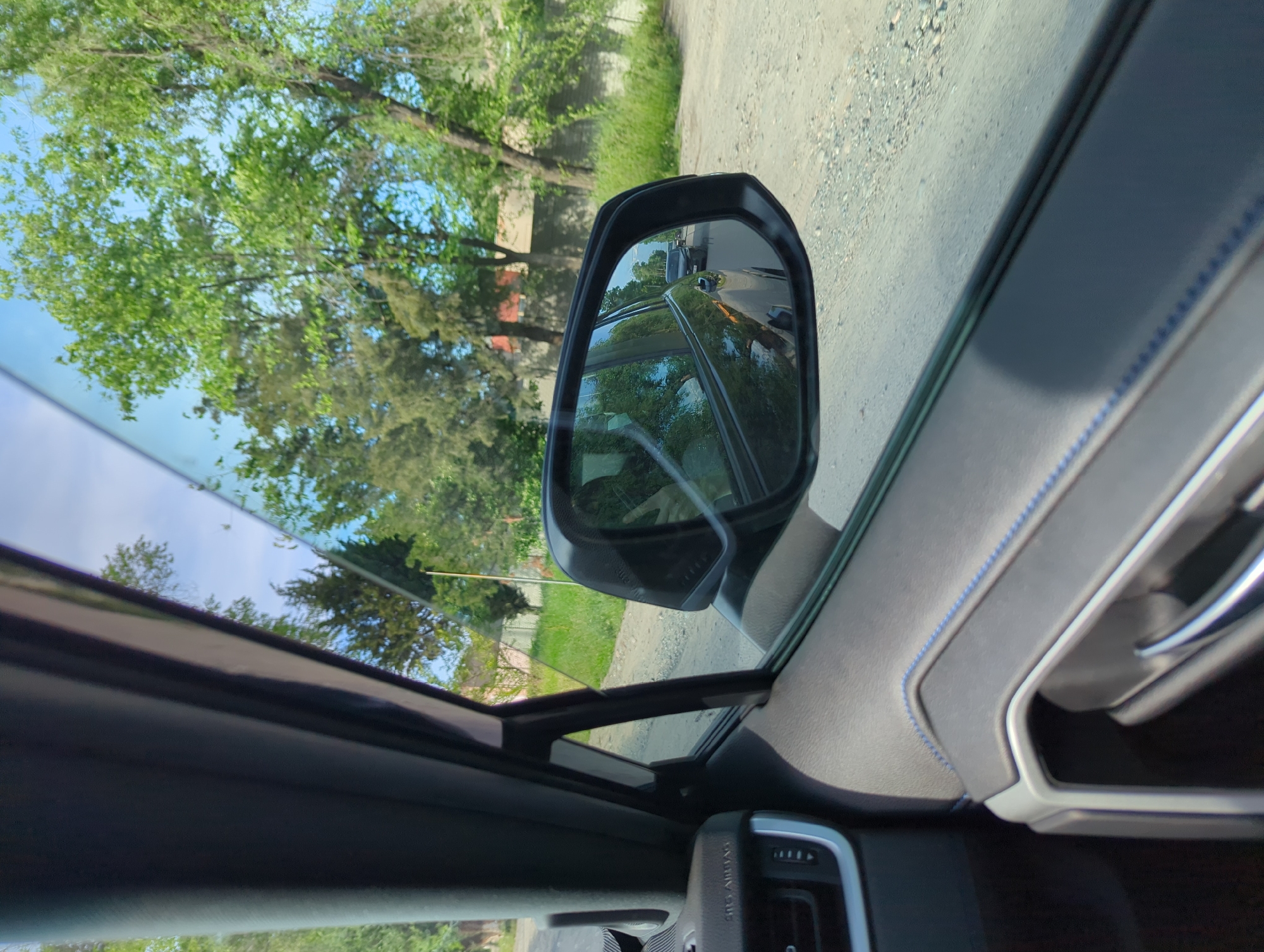 The image shows the interior of a car, focusing on the driver's side window and side mirror. The view through the window includes trees, a blue sky with clouds, and a glimpse of a gravel road. The side mirror reflects the same road and trees, with a vehicle following behind. Inside the car, the door panel is visible, featuring gray upholstery with blue stitching and a silver door handle. A portion of the car's dashboard and controls is also visible. The location is likely Beş-Küŋgöy, Kyrgyzstan, based on the context.