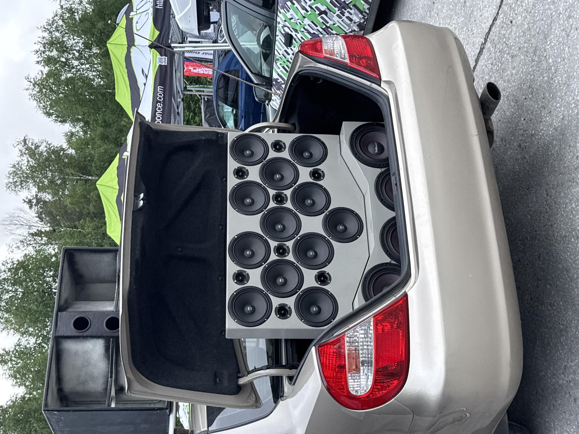 A gold-colored sedan is positioned outdoors on an asphalt surface, its trunk fully open to display a custom-built, multi-component car audio system. The primary speaker enclosure, light gray in color, occupies most of the trunk space and features a grid arrangement of multiple smaller drivers: twelve mid-range speakers and eight smaller tweeters interspersed among them. Below this main unit, a secondary enclosure houses at least three larger subwoofers, one of which has "bewar" branding visible. Perched on top of the open trunk lid is another large, black, rectangular speaker enclosure, which appears to be a horn-loaded design with two prominent circular ports and empty compartments where larger drivers would typically be installed.
In the background, a car audio event is taking place under an overcast sky, with dense green trees forming the horizon. Several event tents are visible; two prominent ones are green, black, and white, with "ponce.com" clearly displayed on one, along with a QR code. A second car, partially visible to the right of the gold sedan, features a distinctive green, black, and white geometric camouflage vinyl wrap, and also appears to have its trunk open. A blue sedan is partially visible further in the background. The presence of the "ponce.com" tent, a known car audio distributor in Russia, indicates the event is likely a car audio competition or show, placing the scene in the city of Perm, Russia. No individuals are visible within the frame.