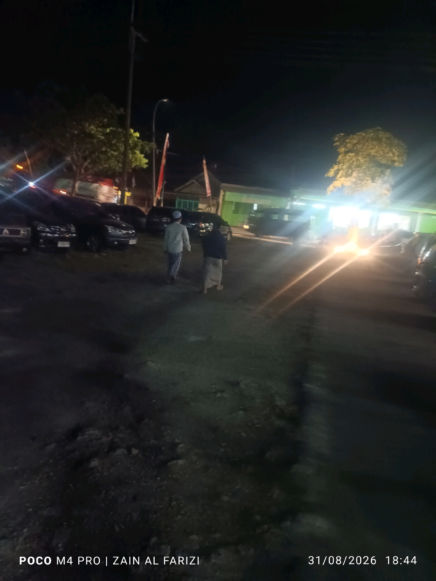 The image depicts a nighttime scene with two individuals walking away from the camera on a paved outdoor area. In the background, several parked cars are visible, along with a building illuminated by artificial lights and trees. The lighting conditions suggest it is after sunset, with artificial light sources casting strong glows and creating lens flare. The overall impression is of a public or semi-public space at night. The text overlay in the bottom left corner indicates the image was taken with a "POCO M4 PRO | ZAIN AL FARIZI", and the bottom right corner shows the date and time "31/08/2026 18:44". While the specific location within Bangkalan, Indonesia is not visually evident from the image itself, it is stated to be in Bangkalan, Indonesia.