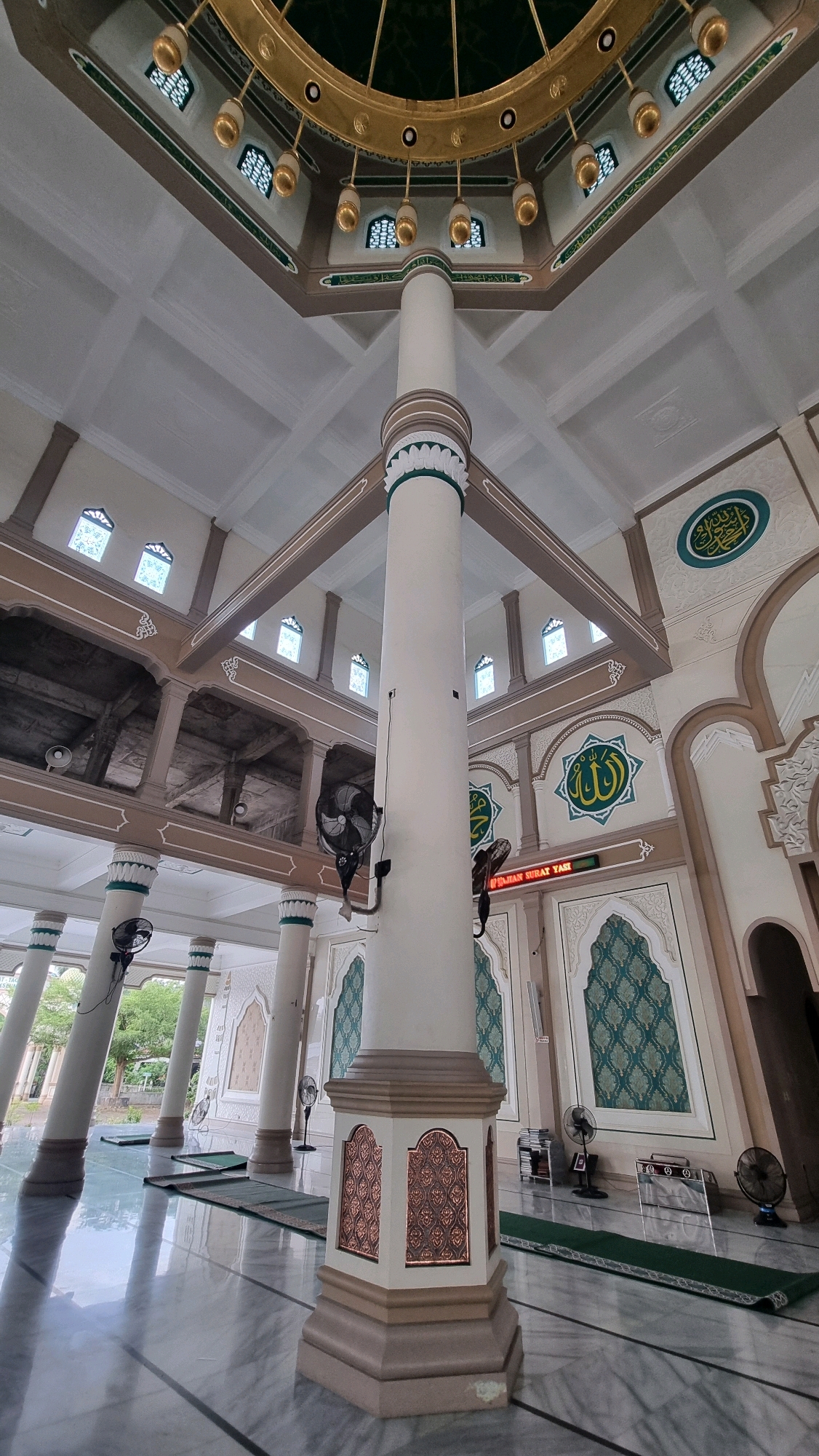 This image captures the interior of the Islamic Center Masjid Agung in Lhokseumawe, Indonesia, offering a low-angle perspective of the grand architecture. A prominent, octagonal white pillar dominates the center of the frame, adorned with decorative reddish-brown panels near its base. Above, the pillar supports a complex ceiling structure with multiple levels and angular beams, all painted in shades of white and beige, accented with green trim. Several small, arched windows with intricate green latticework are visible along the upper walls. Suspended from the golden, circular apex of the ceiling are numerous bell-shaped gold lights. Islamic calligraphy in green adorns the walls, enclosed within circular and arched frames.
The floor is a polished marble with a reflective surface, showing faint patterns of light and shadow. Scattered across the floor are several rolled-up prayer mats in various shades of green, indicating the space is a functional mosque. Wall-mounted black oscillating fans are strategically placed throughout the visible area, with two prominent fans attached to the central pillar and others visible in the background. In the distance, through open arches, glimpses of outdoor greenery can be seen. The overall impression is one of vastness, grandeur, and a serene religious environment.