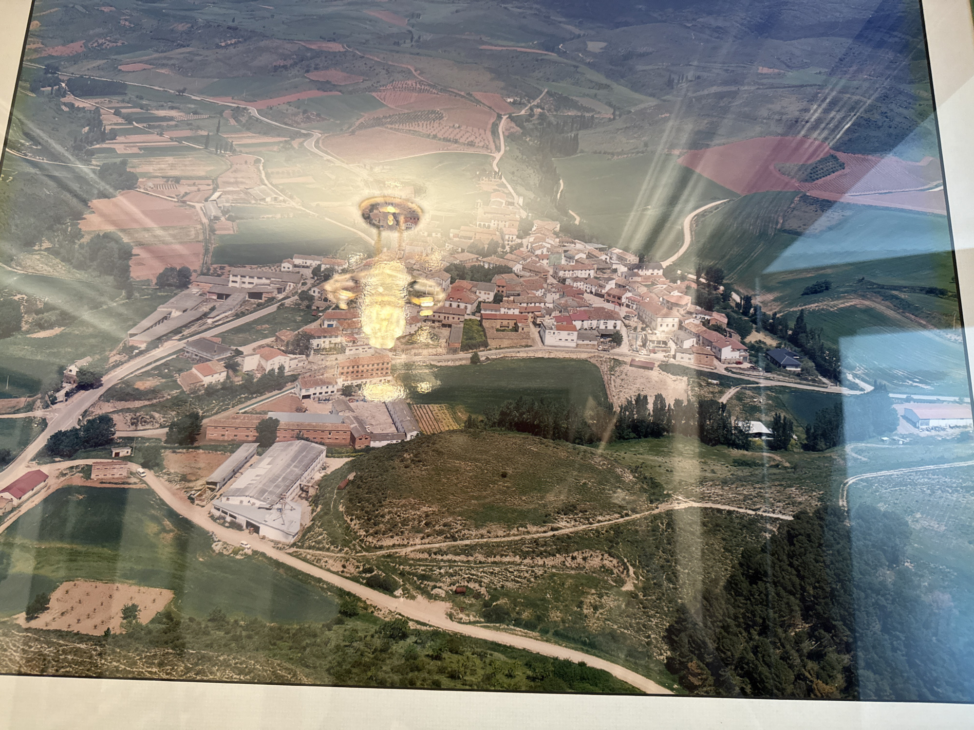 This is an aerial photograph showing a village in the vicinity of Pamplona, Spain. The image depicts a cluster of buildings with red-tiled roofs forming the central part of the village. Surrounding the village are agricultural fields, some of which appear to be cultivated with crops and trees, while others are fallow. Roads wind through the landscape, connecting the village to the surrounding areas. The photograph is taken from a high vantage point, offering a panoramic view of the terrain. There is a notable light flare across the image, particularly in the upper right portion, suggesting it may be a photograph of a screen or a shiny surface. There are no people clearly visible in the image.