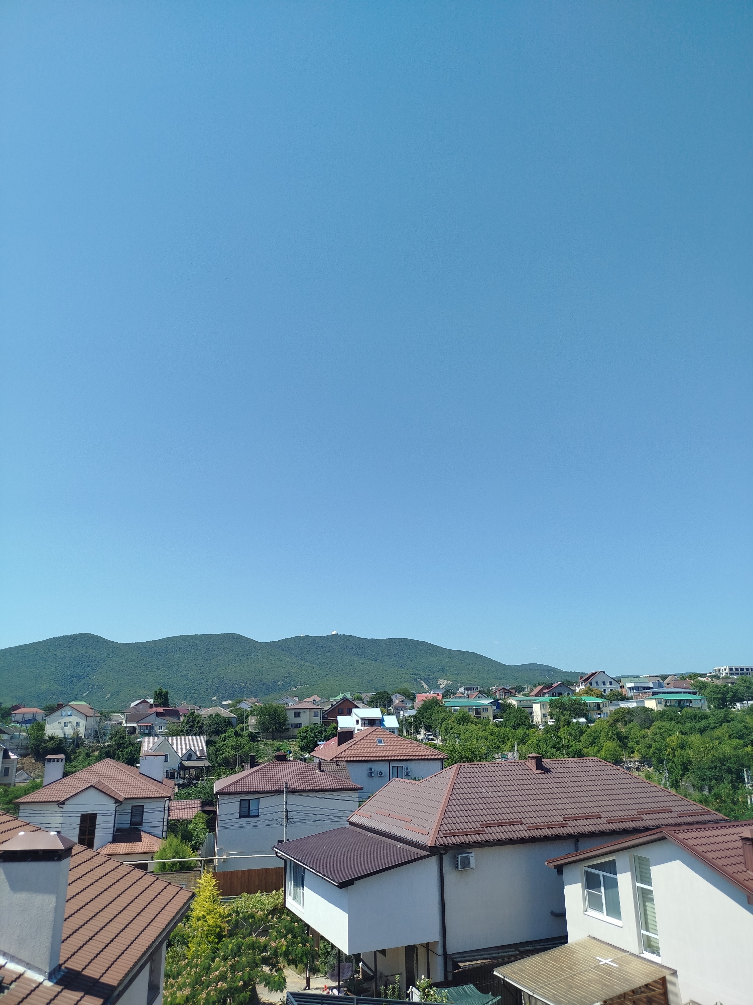The image depicts a view of a residential area in Novorossiysk, Russia, characterized by a cluster of houses with prominent brown tiled roofs. The houses are situated on a gentle slope, with a lush green mountainous backdrop under a clear, bright blue sky. Several trees and vegetation are interspersed among the houses, adding to the suburban feel. The architecture appears to be modern, with multi-story buildings, some featuring balconies and multiple windows. One prominent house in the foreground has a white facade and a large dark brown roof, with a smaller adjacent structure. Another house to its left also has a tiled roof and a chimney. In the distance, the landscape rises into rolling green hills, hinting at the proximity of nature to this developed area. There are no people visible in the scene. The overall impression is one of a peaceful, sunny day in a well-established residential district.
