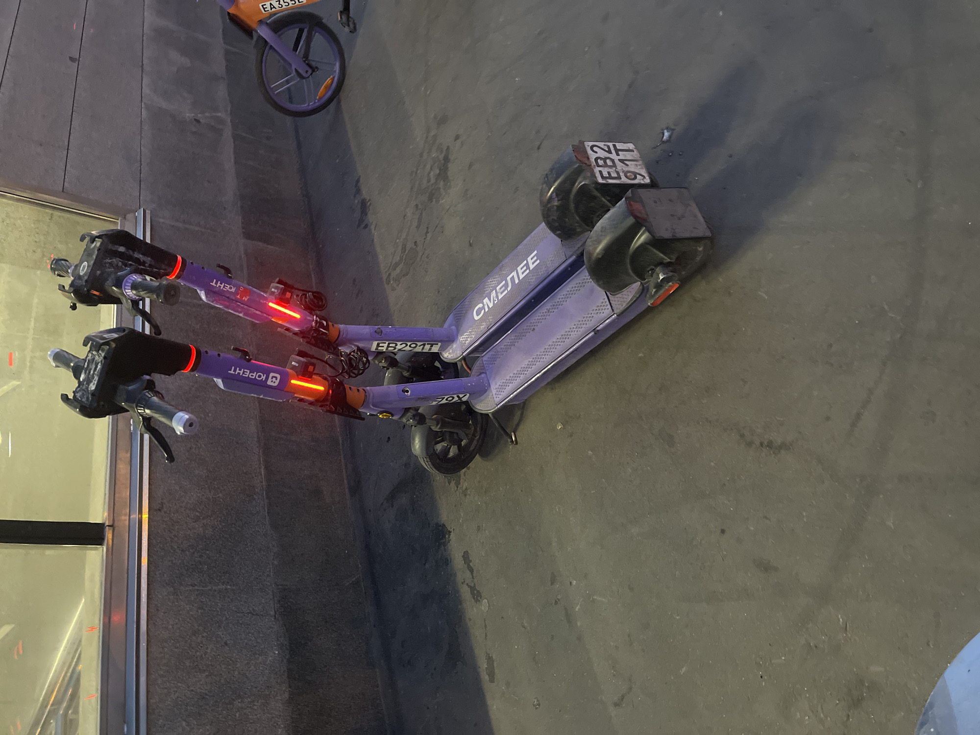 Two purple electric scooters are parked side-by-side on a paved surface. The scooter on the left has the Cyrillic text "ЮПЕРНТ" on its steering column and the text "СМЕЛЕЕ" on its deck. The scooter on the right also has the Cyrillic text "ЮПЕРНТ" visible. Both scooters have illuminated red taillights. The license plate on the rear of the scooter on the left is visible and reads "EB2 91T". In the background, there is a concrete wall with a horizontal band of reflective material and a partially visible orange and purple bicycle. The scene appears to be outdoors, possibly near a public transportation hub or a designated parking area. The lighting suggests it is either dawn, dusk, or nighttime.
