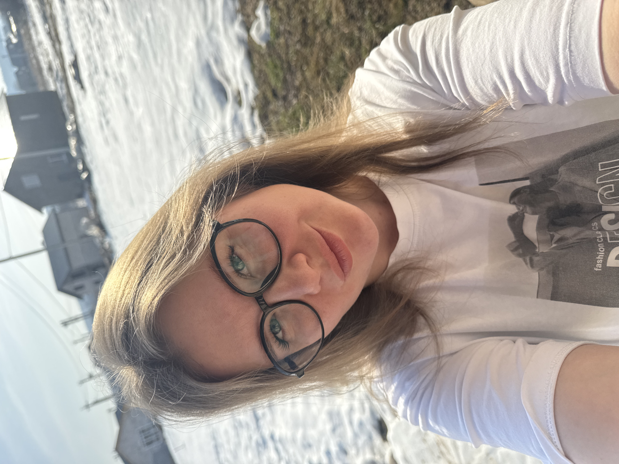 A young woman, wearing black-rimmed round glasses and a white t-shirt, is taking a selfie outdoors. The background shows a snowy landscape with hints of vegetation and buildings, suggesting a location in the vicinity of Gornyi (Dvurechenskoe S/P), Russia. The perspective is close-up and slightly angled, focusing on the woman's face.
