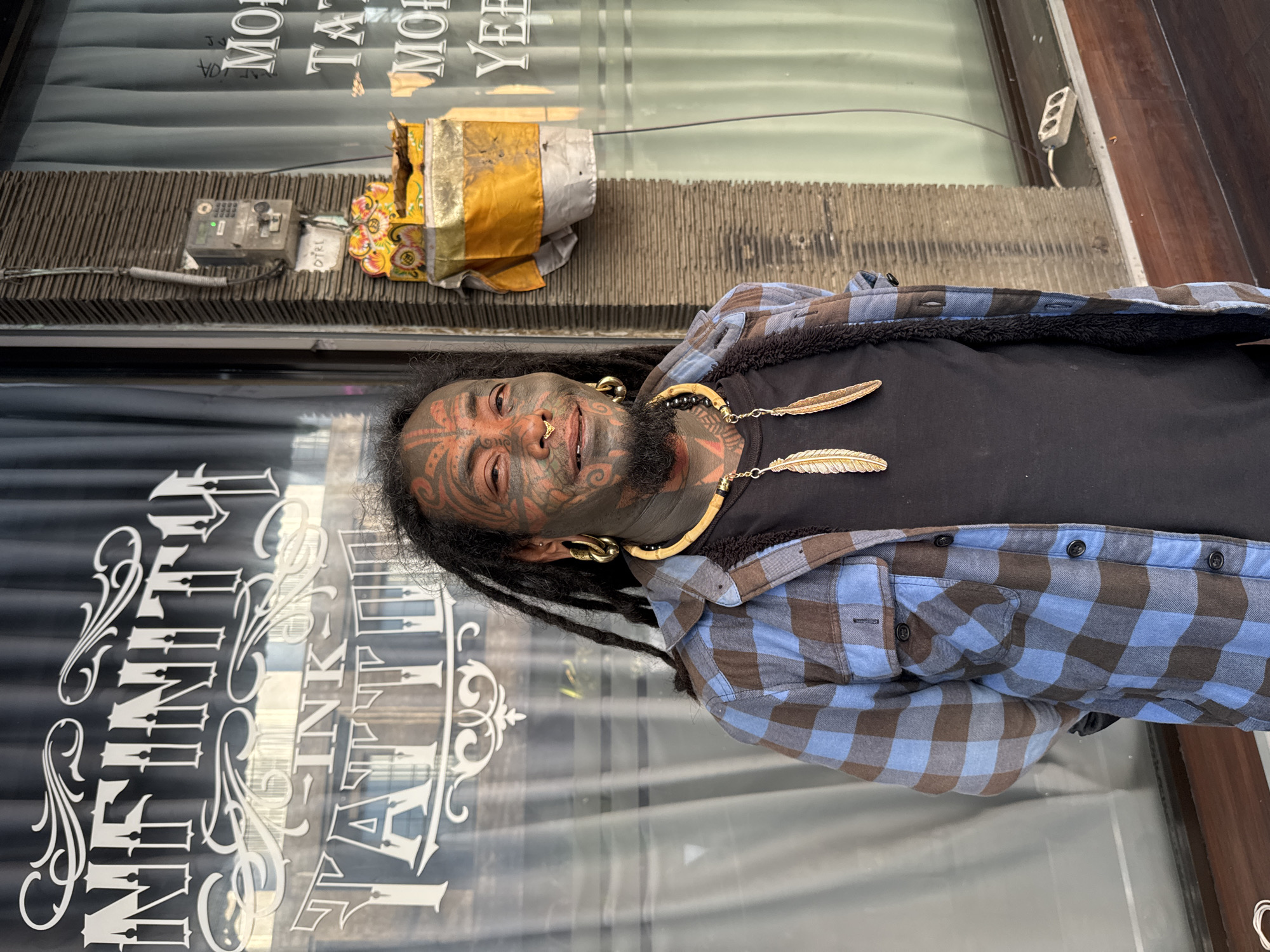A man with extensive facial tattoos, dreadlocks, and large hoop earrings stands outside a tattoo parlor in Kuta, Indonesia. He wears a blue and brown plaid shirt over a black t-shirt and a beaded necklace with two feather pendants. The tattoo parlor's window displays the words "INFINITY INK TATTOO" in white lettering. To the right of the man, a concrete pillar holds a small, colorful shrine and a wall-mounted electrical box. The ground is a dark wooden plank.