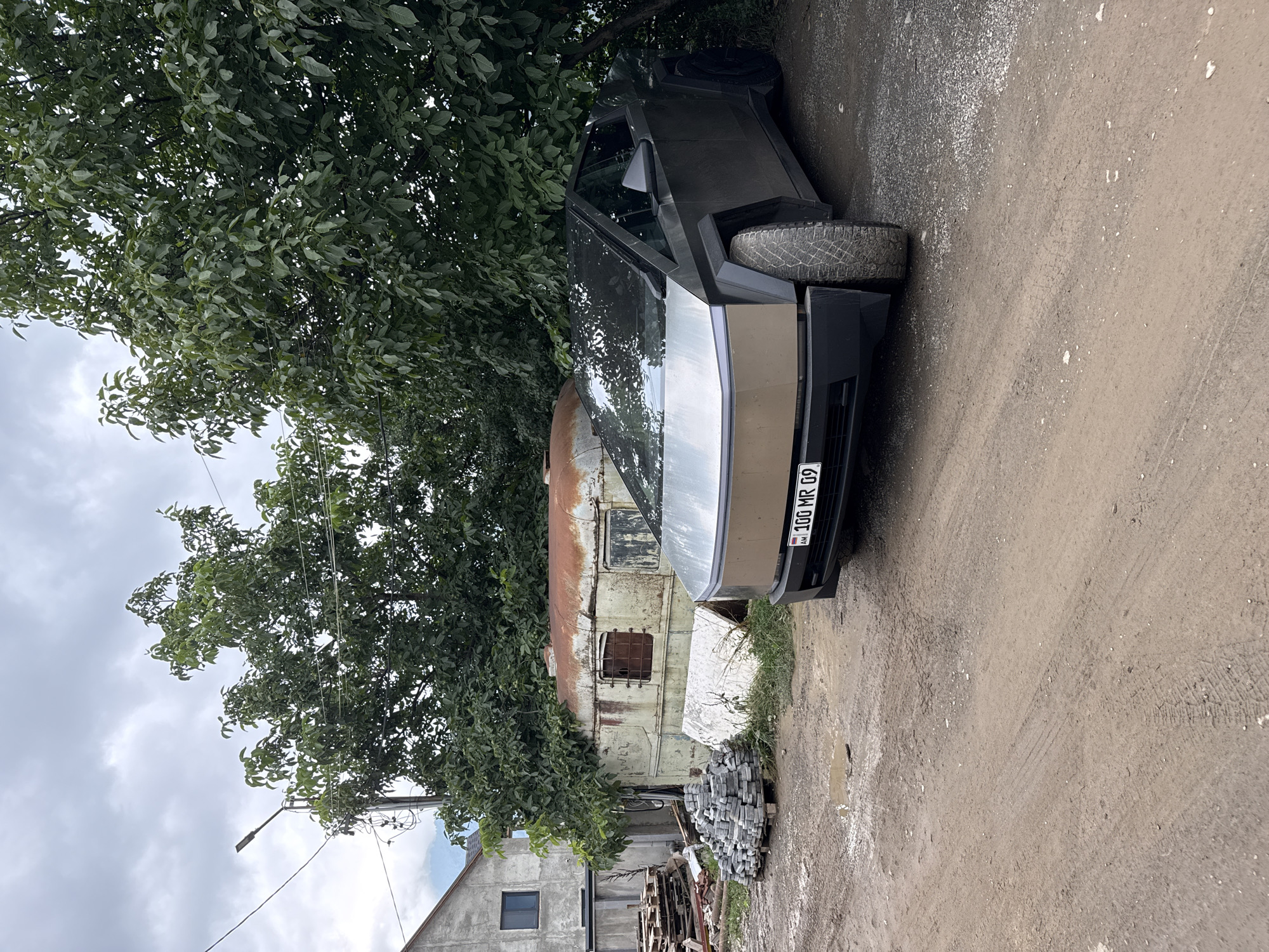 The image depicts a futuristic-looking vehicle, possibly a Tesla Cybertruck, parked on a dirt road in the city of Dilijan, Armenia. The vehicle is dark gray and has a distinctive angular design. It is positioned to the right of a dilapidated, rusty metal structure that appears to be an old trailer or shed. In the background, a building with a light-colored facade and a dark window is visible. To the left of the trailer, there is a pile of grey paving stones stacked on wooden pallets. A large tree with lush green foliage dominates the upper portion of the image, casting shadows on the scene. The ground is uneven and muddy, with tire tracks visible, suggesting recent vehicle activity. The sky is overcast and cloudy. The license plate of the vehicle reads "AM 100 MR 09".