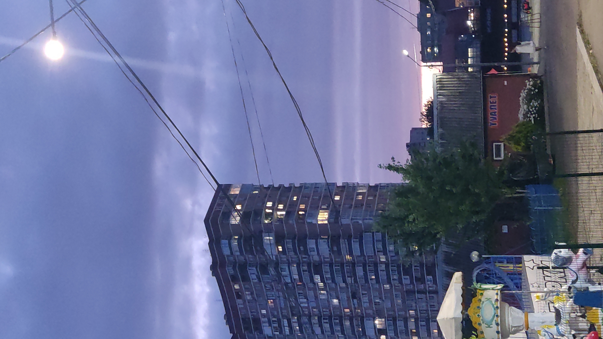 This is a vertical, eye-level, outdoor, full shot of a city scene at dusk. In the foreground, a small carousel with white and yellow striped canopy is visible to the left. Beyond the carousel, a tall, brick residential building with many lit windows dominates the left side of the frame. In the middle ground, a small, red building with a sign that reads "ТУАЛЕТ" (TOILET) in Cyrillic lettering is visible, with a tree and some bushes in front of it. In the background, the sky is dark and cloudy, with a pale sunset visible on the horizon. There are several power lines stretching across the frame. A street lamp is visible in the upper right quadrant of the image, emitting a bright light. A person in a white dress is walking on a paved area to the right of the red building. The scene is likely a park or recreational area within the city of Krasnodar, Russia, given the Cyrillic signage.