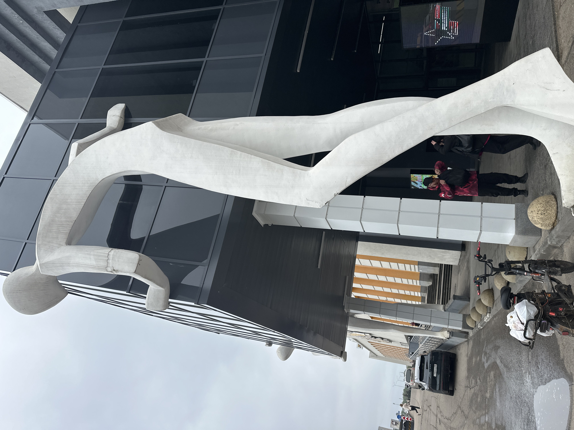 The image shows a modern sculpture of an abstract human figure in front of a glass-faced building. The sculpture is white and appears to be made of stone or concrete. Two people are standing underneath a portion of the sculpture near the entrance of the building. There is a bicycle and a car parked nearby on the street. The location is Nizhny Novgorod, Russia.